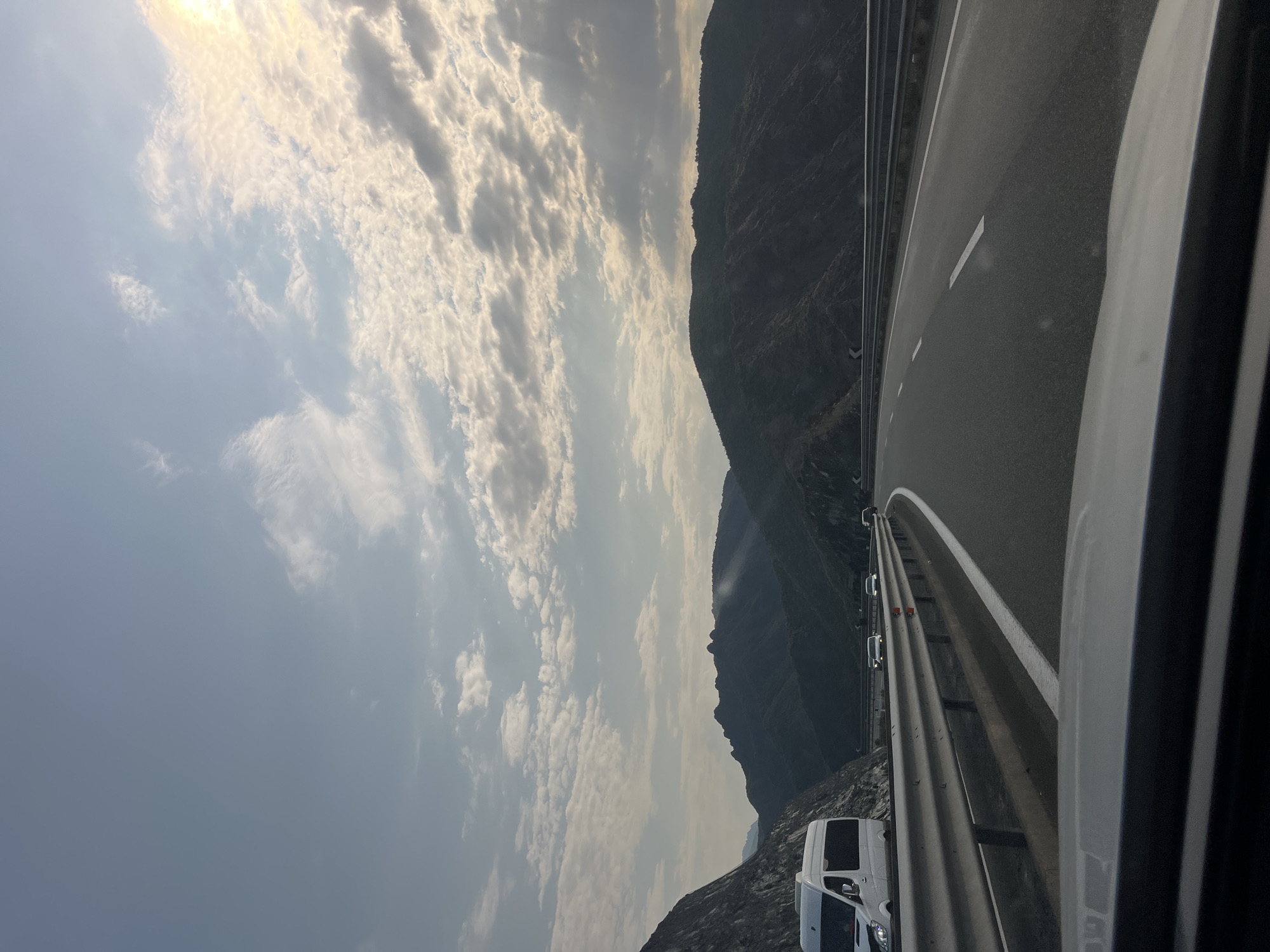 The image depicts a highway in Rrëshen, Albania, at dusk. The road curves to the right, bordered by a metal guardrail. To the left, a white van is parked on the shoulder. Mountains dominate the background, shrouded in shadow. The sky is filled with clouds, illuminated by the setting sun, which casts a warm glow. The overall scene conveys a sense of travel and the vastness of the natural landscape.