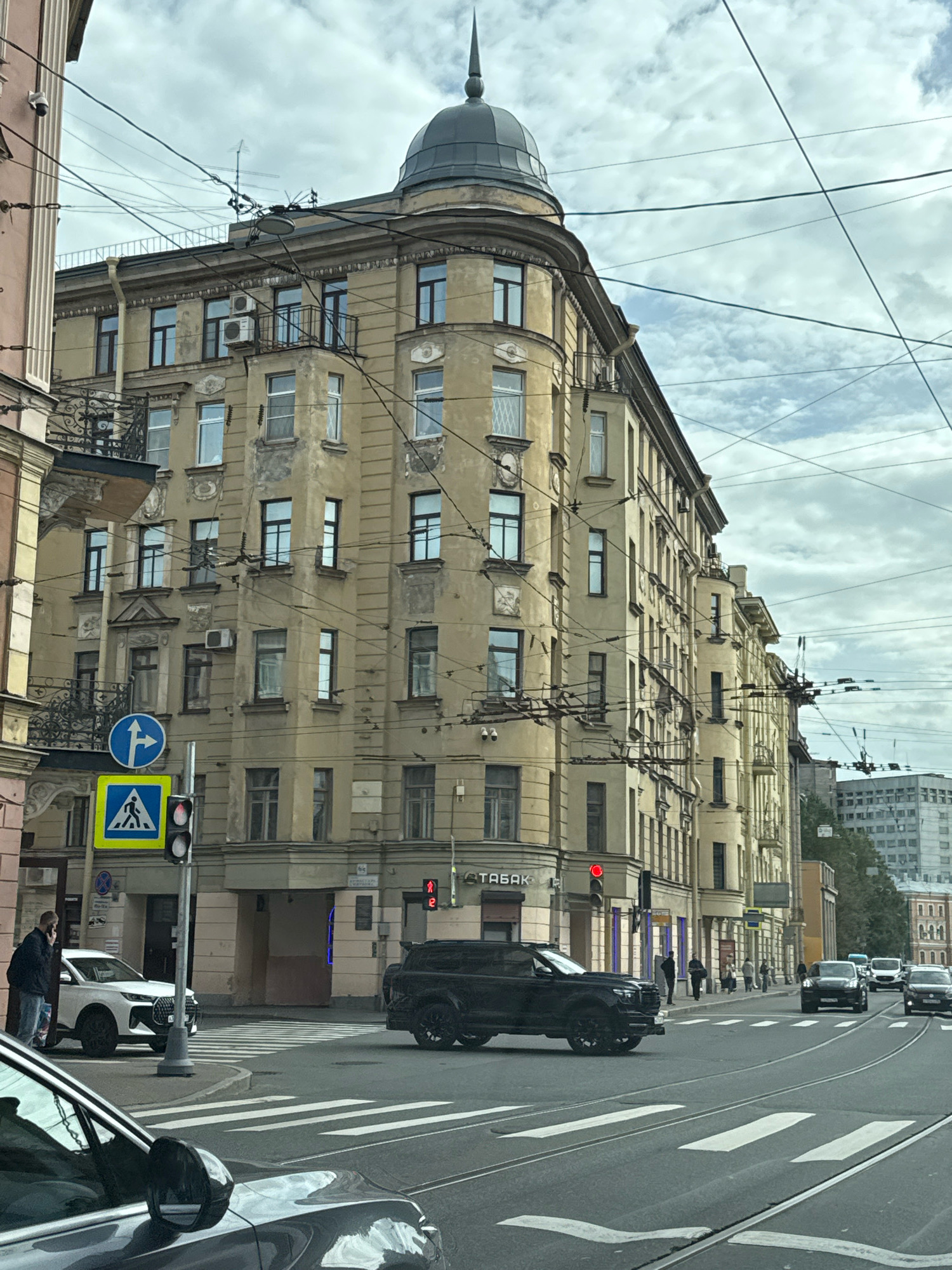 A street-level, eye-level, medium shot captures a cloudy day in Saint Petersburg, Russia, focusing on the corner of a weathered, multi-story building with a distinctive domed turret. The building, likely dating back to the early 20th century, features ornate architectural details, including decorative moldings and balconies with intricate wrought-iron railings. The facade is a pale yellow, showing signs of age and weathering. Overhead, a complex network of electrical wires for trams crisscrosses the sky, typical of the city's infrastructure.
In the foreground, a black SUV is stopped at a crosswalk, its rear facing the camera. To the left, a white SUV is also stopped at the crosswalk, with a person in a dark jacket and jeans standing next to it, talking on a mobile phone. Traffic lights indicate red, halting vehicles. A pedestrian crossing sign and a directional sign are visible on a pole near the traffic light. Across the street, several other cars are visible in traffic, and pedestrians can be seen walking on the sidewalk in front of shops. The street is paved with asphalt and marked with white pedestrian crossings and tram tracks. The background reveals more buildings, including a modern, grey high-rise structure on the right, contrasting with the older architecture. The overall atmosphere is urban and bustling, with a sense of everyday city life unfolding under a cloudy sky.