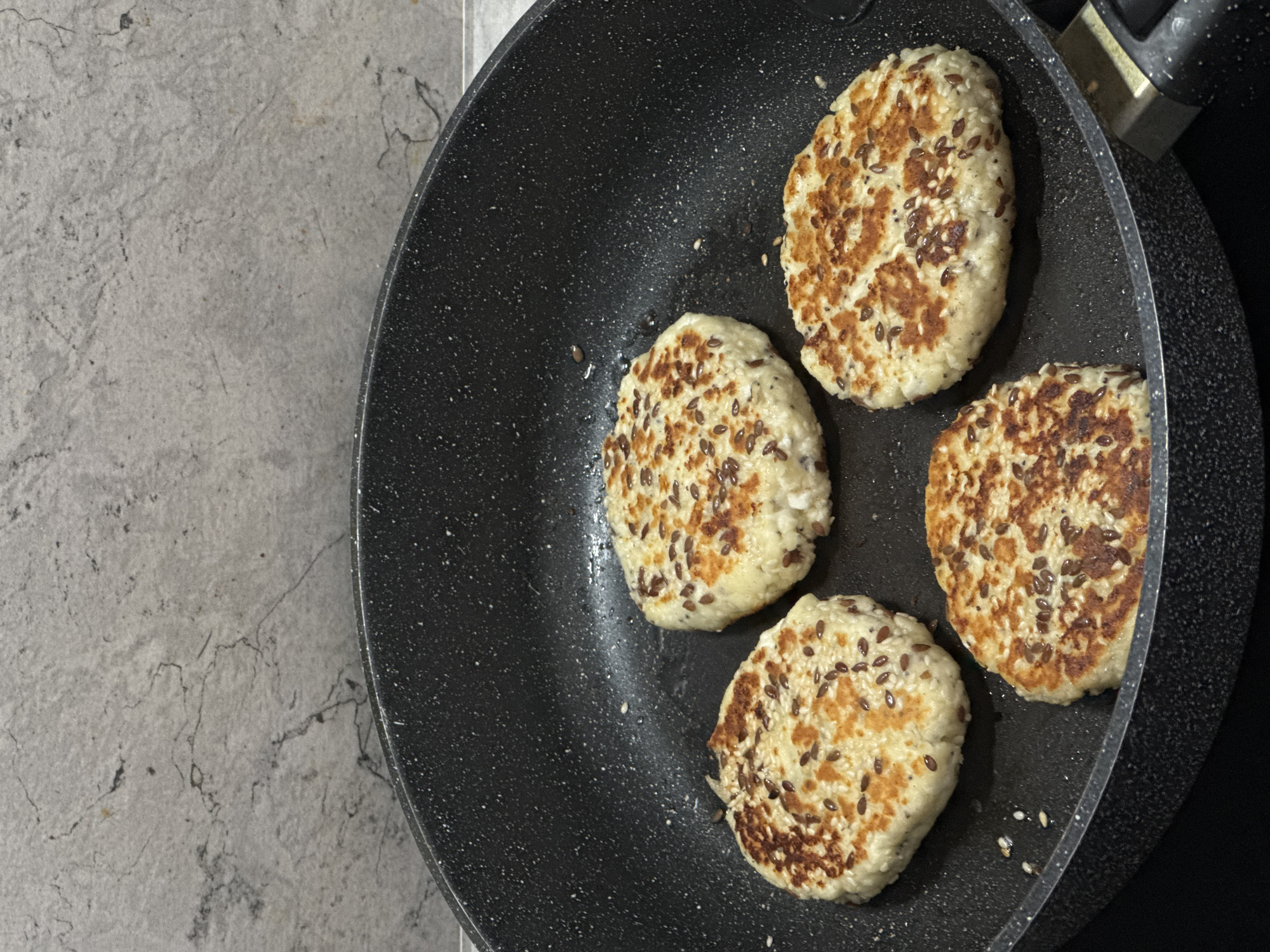 Four round, golden-brown patties with visible seeds, likely sesame and flax, are cooking in a black non-stick frying pan. The pan is placed on a dark stovetop, and the background features a grey, speckled wall, resembling a kitchen backsplash. The patties appear to be made of a light-colored mixture, possibly a dough or batter, and are evenly browned on their visible sides, suggesting they are in the process of being fried or pan-seared. The lighting is neutral, casting minimal shadows and highlighting the texture of the patties and the speckled pattern of the pan and wall. No people are visible in the image, and the location is not specified beyond being a kitchen environment.