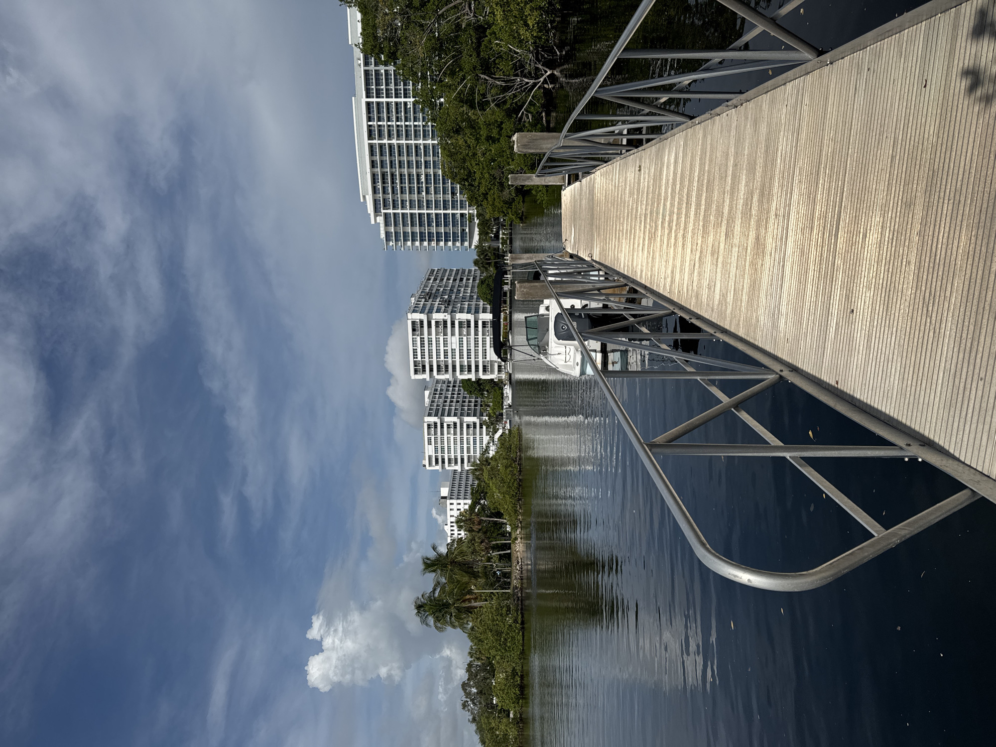 The image depicts a bright, sunny day with a partly cloudy sky over a body of water. In the foreground, a wooden pier with metal railings extends into the water. A small white boat is docked to the left of the pier. Across the water, lush green trees and vegetation line the shore, with several tall, modern white apartment buildings situated behind them. The buildings feature numerous balconies and windows, and their reflections are visible on the calm water. The scene appears to be a waterfront residential area in Fort Lauderdale, Florida, likely near the Intracoastal Waterway or a similar canal system, given the presence of the apartment buildings and boat docks typical of the city.