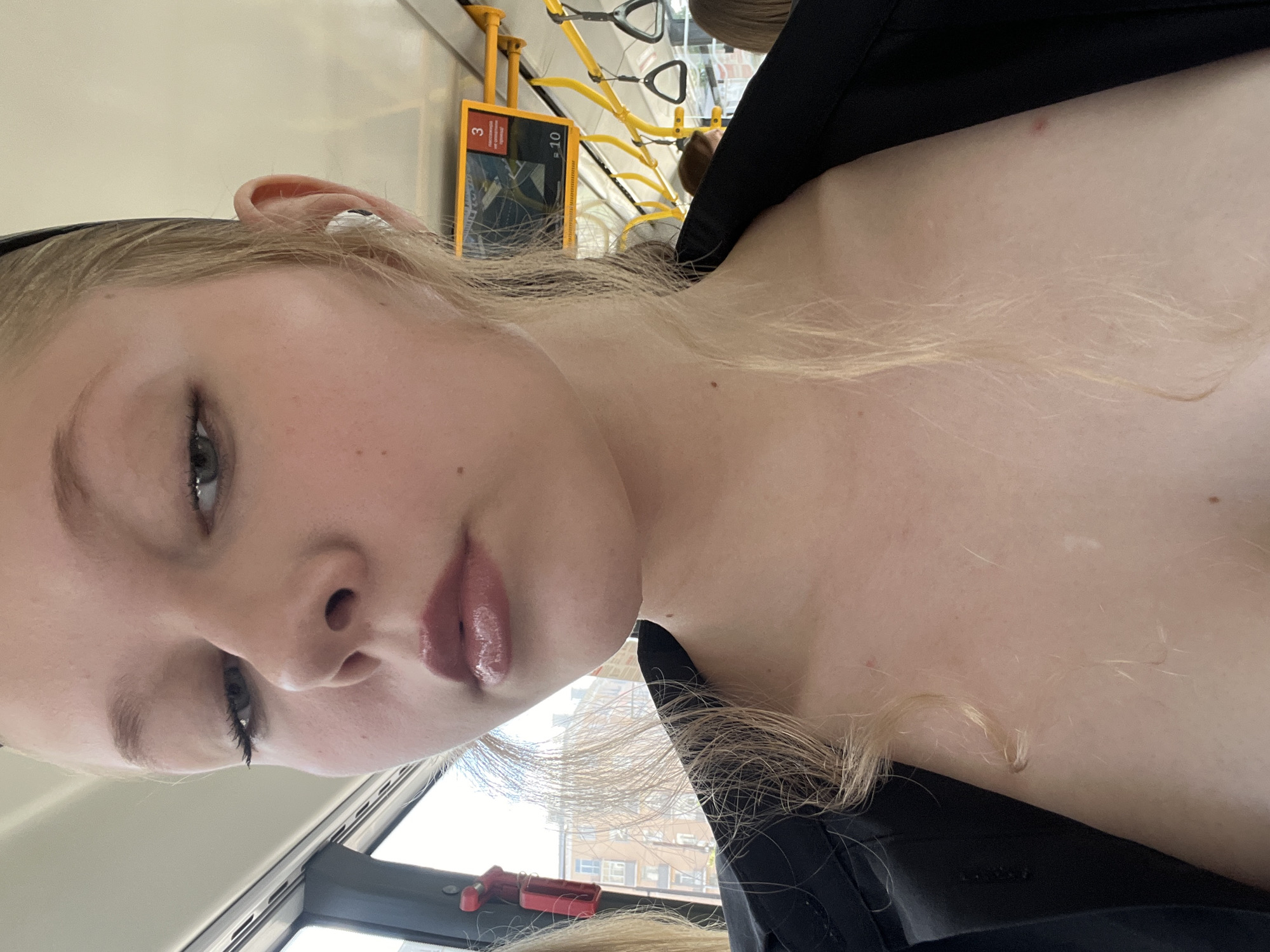 A close-up shot features a young woman with blonde hair and fair skin, wearing a black garment. She has light makeup on, including lip gloss, and is wearing white earbuds. Her hair is styled in loose waves, with some strands falling forward. The background suggests she is inside a public transportation vehicle, possibly a bus or tram, with yellow poles and straps visible. A sign with the number "3" and some text in Russian is mounted above. The environment appears to be urban, with buildings visible through the window. The overall lighting is natural and bright.