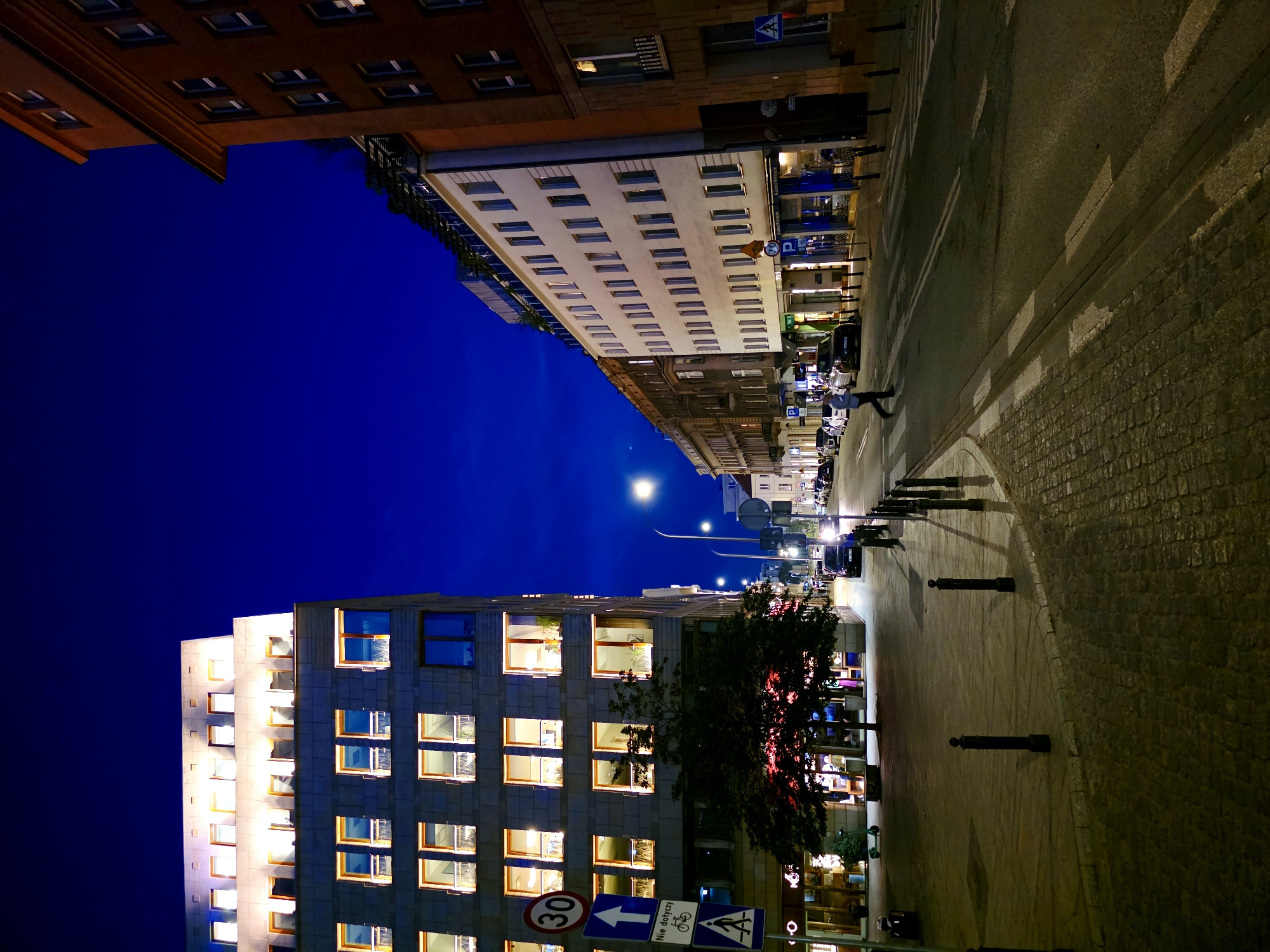 The image depicts a city street in Warsaw, Poland, at night, likely during twilight as the sky is a deep blue. On the left, a modern, illuminated building with multiple floors of lit windows stands prominently. In front of this building, a sidewalk is paved with cobblestones, and bollards line the edge of the street. A tree with red-lit foliage is situated on the sidewalk. Several street signs are visible, including a "30" speed limit sign and a pedestrian crossing sign with Polish text. A crosswalk is marked on the street.
To the right of the modern building, an older, multi-story building with a brick facade is visible. Further down the street, more buildings are seen, including a lighter-colored building with numerous windows. Cars are parked along the street and some are in motion. A lone pedestrian is crossing the street at the crosswalk. Streetlights illuminate sections of the road and sidewalk. The overall impression is of a well-lit urban environment after dark.