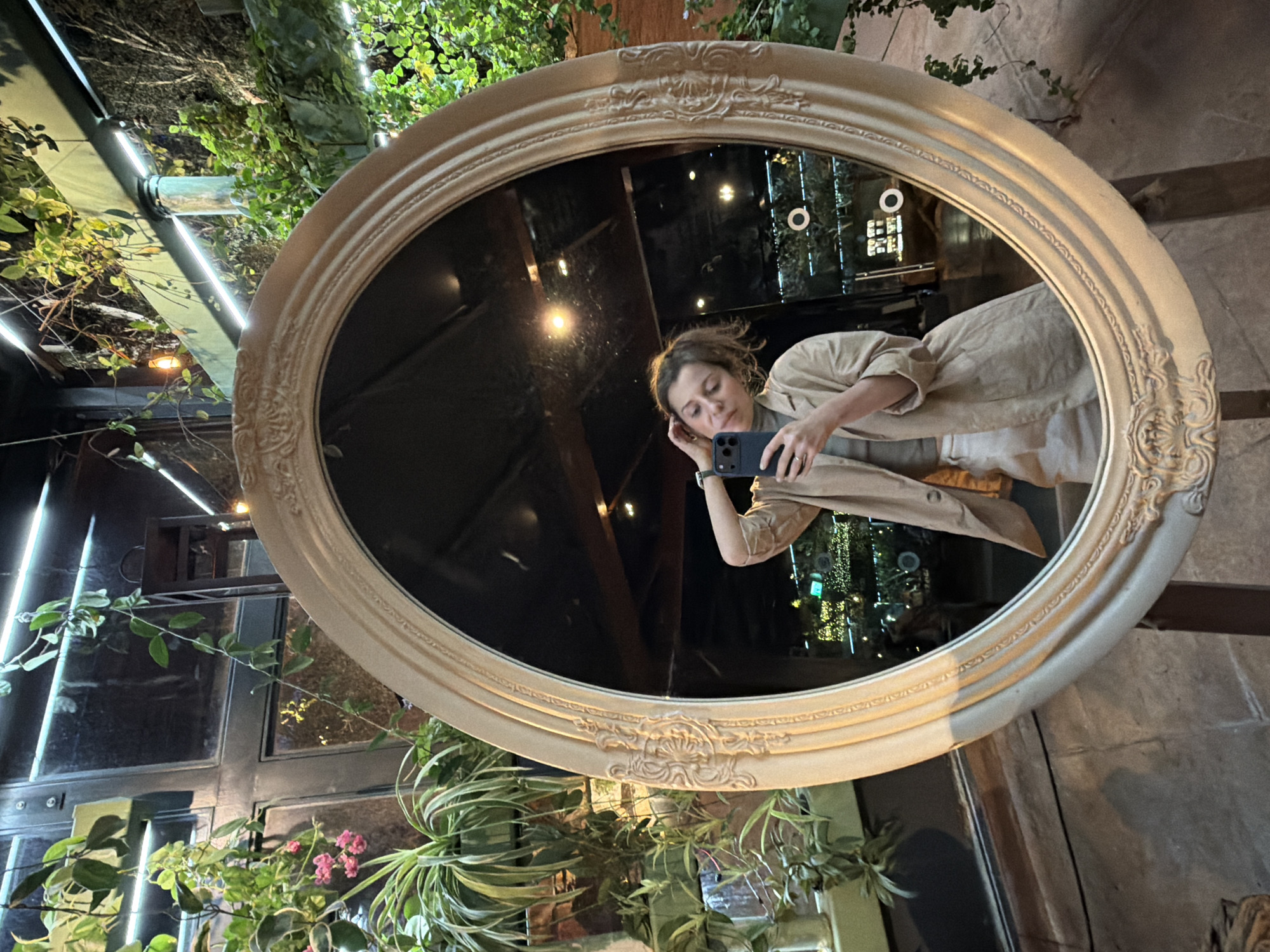 This image captures a woman taking a mirror selfie in a lush, green-filled indoor or semi-enclosed setting, likely during the evening or night in Nir Eliyahu, Israel.
The central object is a large, oval mirror featuring an ornate, light-colored frame. Reflected in the mirror is a woman with light brown hair, dressed in a light-colored, possibly beige or khaki, top or jacket. She holds a dark-colored smartphone, resembling an iPhone, in her right hand, angled to photograph her reflection. Her left hand is casually placed near her head. Her expression is calm and focused on the task of taking the picture.
The immediate surroundings outside the mirror are rich with various green plants and vines, some climbing on structures. Bright fluorescent strip lights are visible on what appears to be a ceiling beam or canopy, casting light on the foliage. A textured, possibly stone or concrete, wall is also visible near the mirror.
Within the mirror's reflection, behind the woman, more artificial lighting is evident, including warm-toned lights that could be fairy lights or small bulbs, contributing to a cozy and atmospheric ambiance. The overall lighting is dim, highlighting the prominent artificial light sources. No visible text is present in the image.