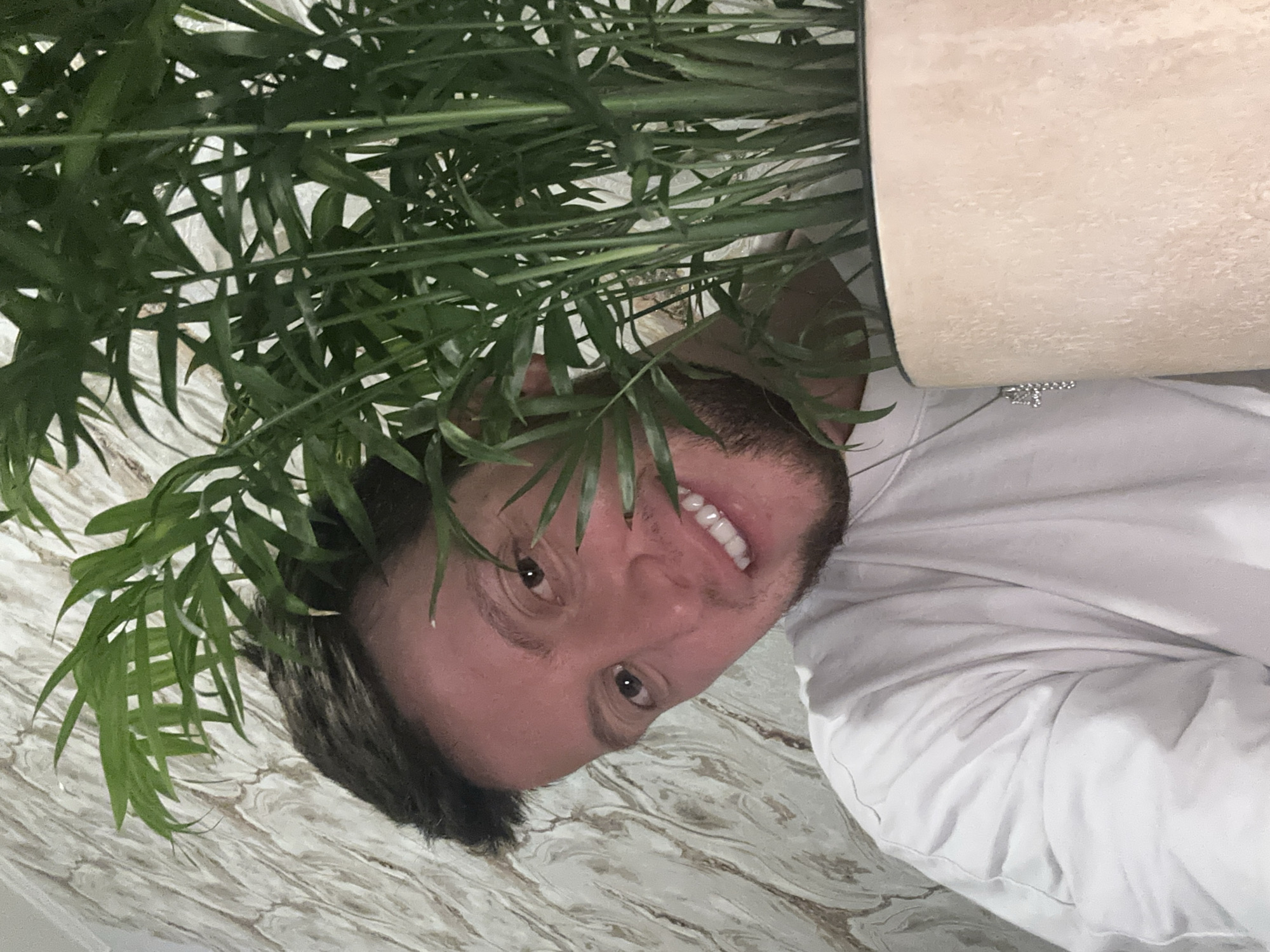 A close-up, waist-up portrait shows a young man with dark hair and brown eyes peeking out from behind a potted houseplant. He is wearing a white t-shirt with a silver cross necklace. The background is a textured, light-colored wallpaper with a marble-like pattern. The man is smiling and looking at the camera. The lighting is soft and natural. The image appears to have been taken indoors. The man is positioned to the left of the frame, with the plant dominating the right side.