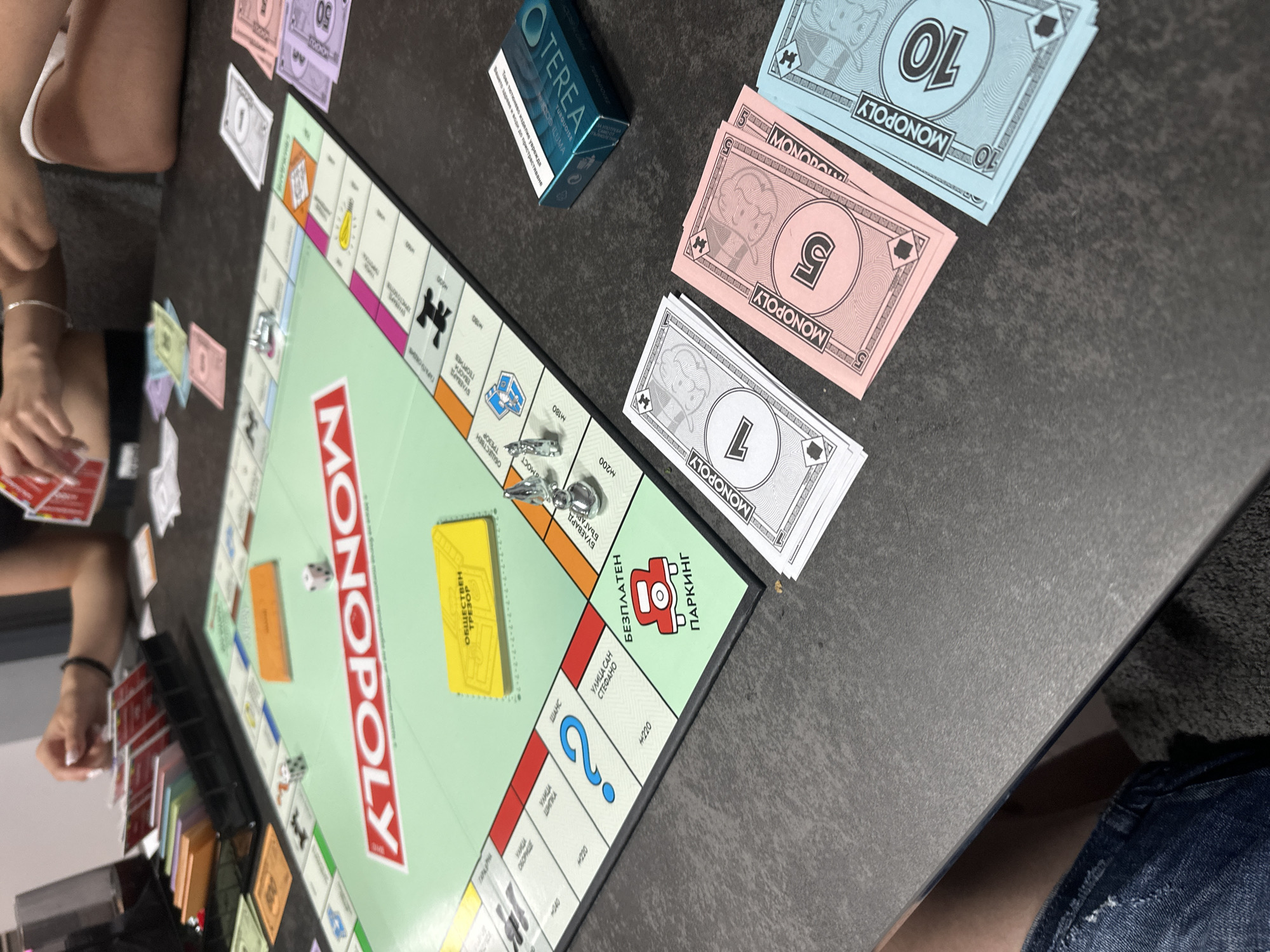 The image depicts a top-down view of a Monopoly board game in progress on a dark, textured table. The game is set in an indoor environment, likely a home or game room, with no specific city or location identifiable. Several hands are visible, interacting with the game. One hand, with painted fingernails, is holding red Monopoly cards. Another hand, partially visible, is holding more cards.
The Monopoly board is predominantly green with red and white lettering spelling out "MONOPOLY." Several game pieces are on the board, including two dice, one white and one black with white pips, showing a 2 and a 4 respectively. There are also two silver-colored game tokens, one resembling a thimble and the other a small car. A yellow "COMMUNITY CHEST" card is placed on the board, and a "FREE PARKING" space with a red car icon is visible. Scattered around the board are Monopoly money bills in denominations of 1 (white), 5 (pink), and 10 (blue). A teal-colored box, identified by the text "TEREA TURBOKOIZE," is placed to the right of the board. The background shows a glimpse of furniture, possibly chairs. The scene suggests an active game of Monopoly with players engaged in gameplay.