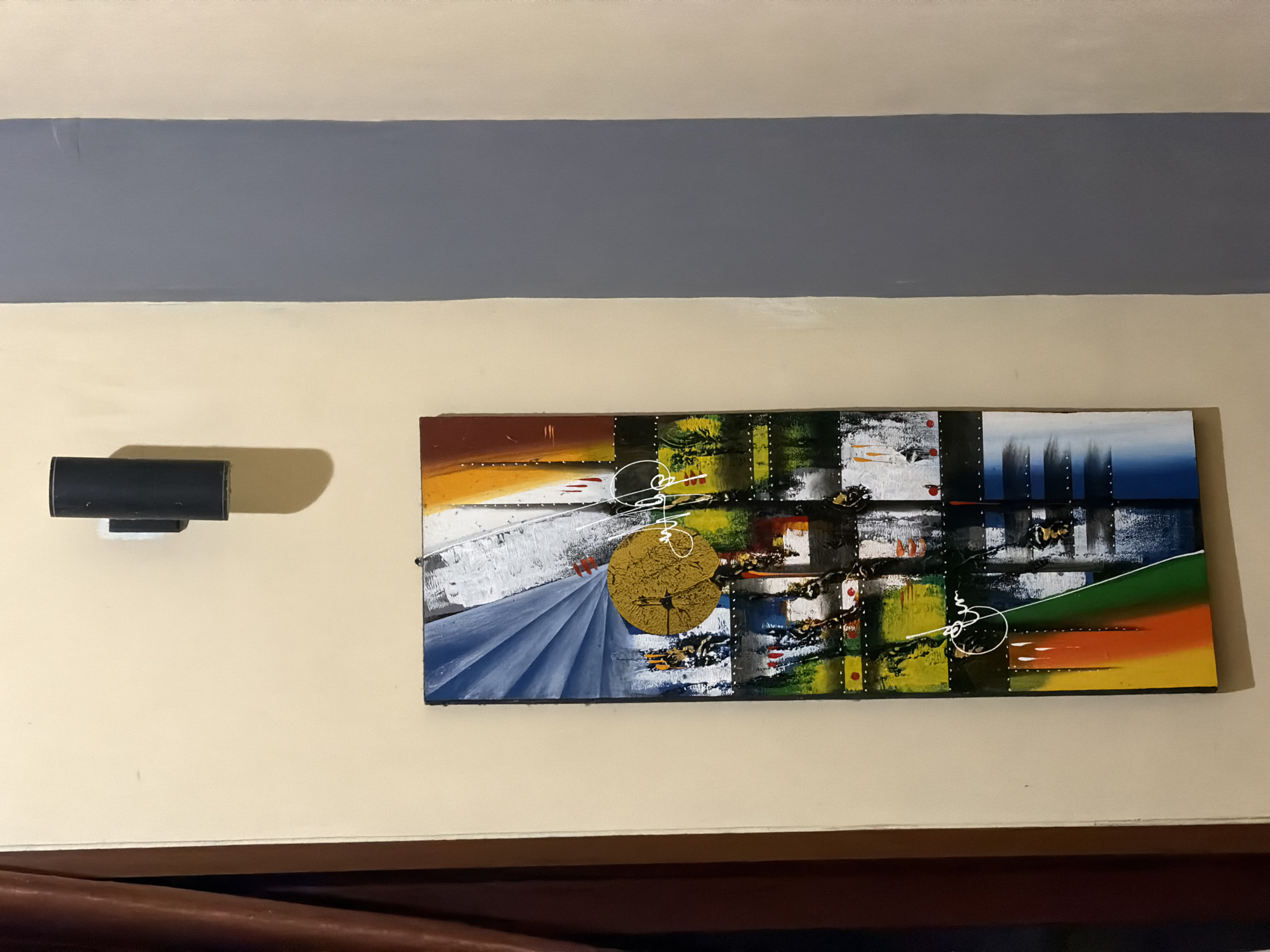 The image shows a vertical, abstract painting with bold colors and geometric shapes hanging on a cream-colored wall. Above the painting, a black cylindrical light fixture is mounted on the wall. To the right of the cream wall, a vertical strip of gray painted wall is visible. The painting features a circular yellow element in the upper half, surrounded by abstract brushstrokes in blue, white, orange, and brown. The lower half of the painting includes sections of green, black, blue, and a gradient of yellow, orange, and red. White, linear elements are also incorporated into the abstract design. The overall impression is of a modern, artistic composition. The context of Wonoayu, Indonesia, is not visually discernible from the image itself.