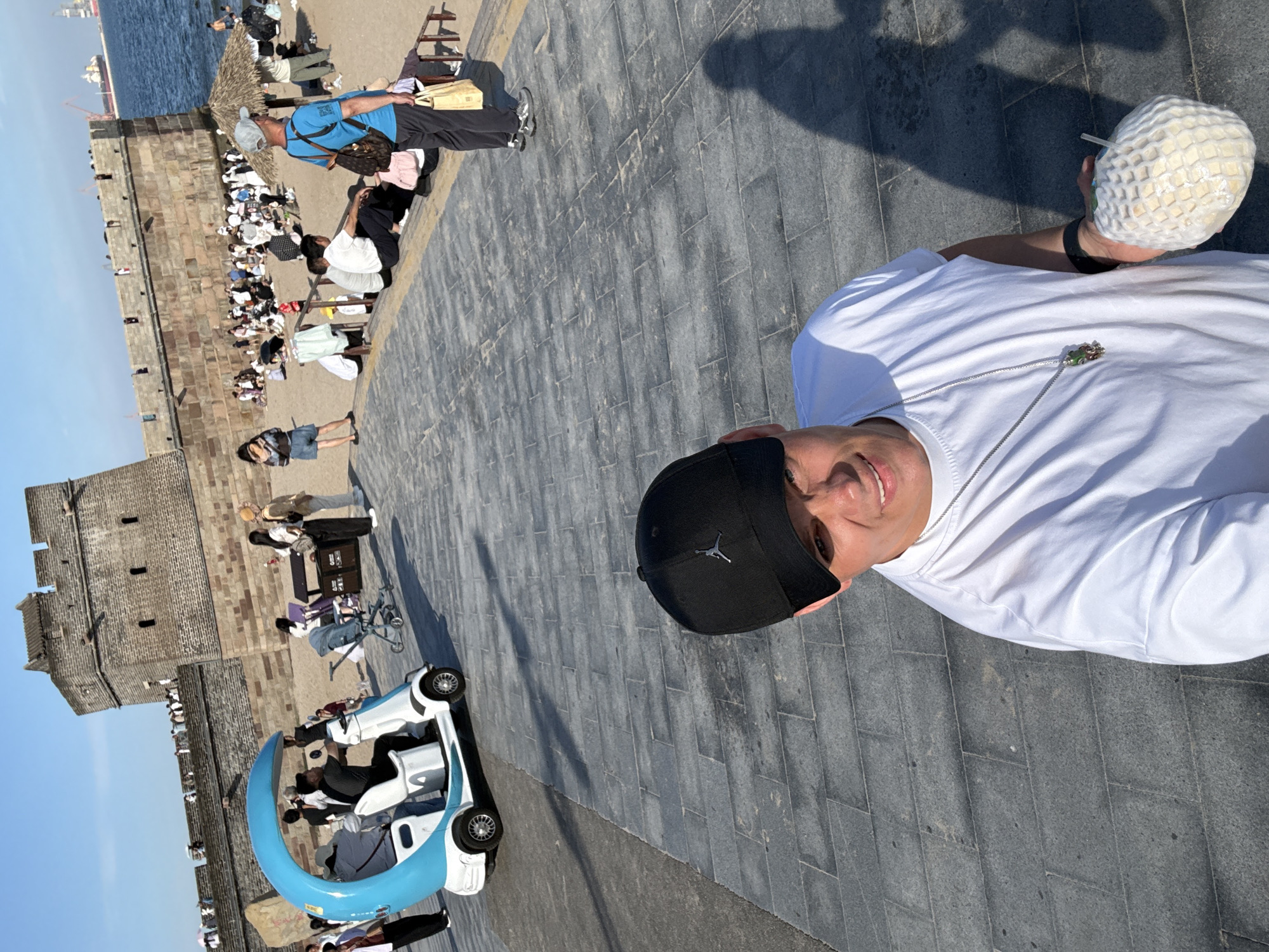 This is a wide-angle, eye-level selfie taken outdoors in Qinhuangdao, China, on a sunny day. The foreground features a man wearing a black baseball cap and a white t-shirt, holding a netted coconut in his right hand. He is smiling and looking towards the camera. In the background, a section of the Great Wall of China, a historic stone fortification, is visible, overlooking a sandy beach and the sea. Numerous tourists are scattered across the beach and along the wall, some sitting, others standing, and some walking. A distinctive blue and white mini-electric vehicle, resembling a small golf cart, is parked on a paved walkway in the mid-ground. The walkway is made of grey stone pavers, and the beach is golden sand. The sky is clear and blue, with a few distant cranes visible near the horizon, indicating port activity. The overall scene depicts a popular tourist destination where historical architecture meets recreational beach activities.