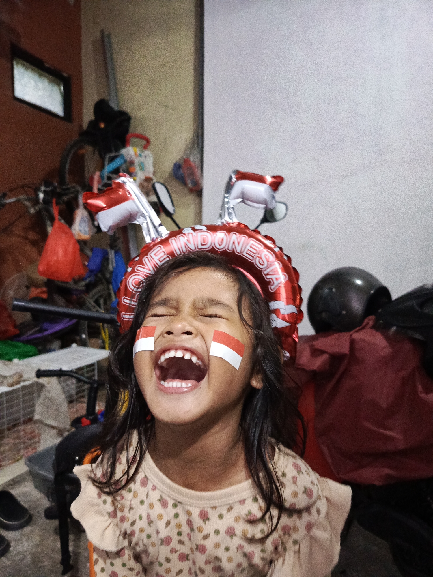 A young girl with her eyes closed and mouth open in a wide, joyful laugh is pictured in what appears to be a garage or storage area in Ciseeng, Indonesia. She wears face paint in the Indonesian flag colors of red and white on her cheeks, and a red, inflatable headband that spells out "LOVE INDONESIA" in white lettering. Red and white balloons, also representing the Indonesian flag, are positioned above her head. The background shows bicycles, shelves with various items, and a motorcycle with a black helmet. The overall environment suggests a domestic or semi-commercial space, possibly decorated for a national holiday or celebration.
