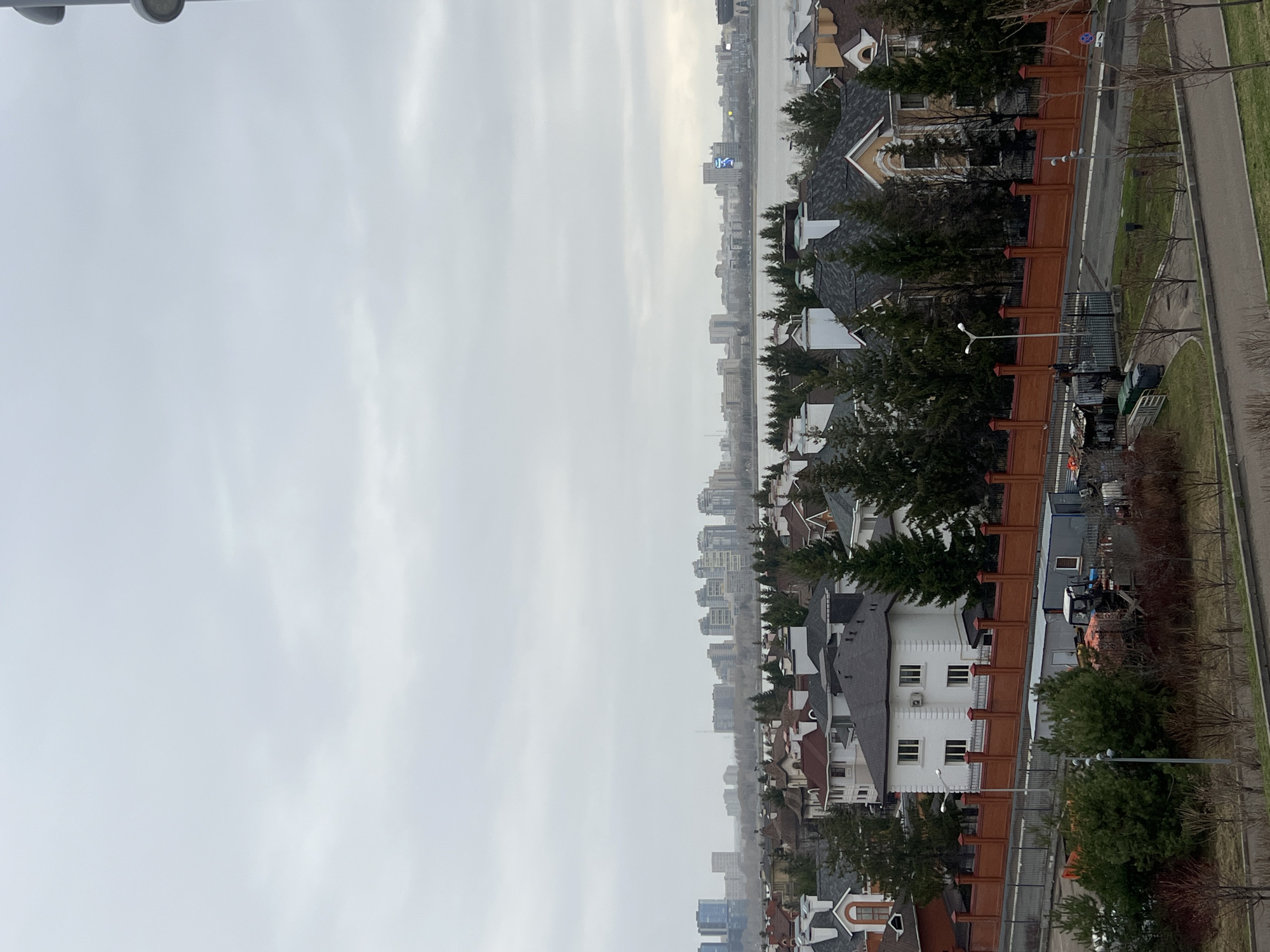The image shows an elevated view of a residential area in Kazan, Russia, under an overcast sky. The foreground features a row of houses with varying architectural styles, including structures with white facades, dark grey roofs, and brown fencing. Lush green trees are interspersed among the houses and along the street. A paved walkway runs parallel to the road. The distant cityscape reveals taller buildings partially obscured by the hazy atmosphere.
