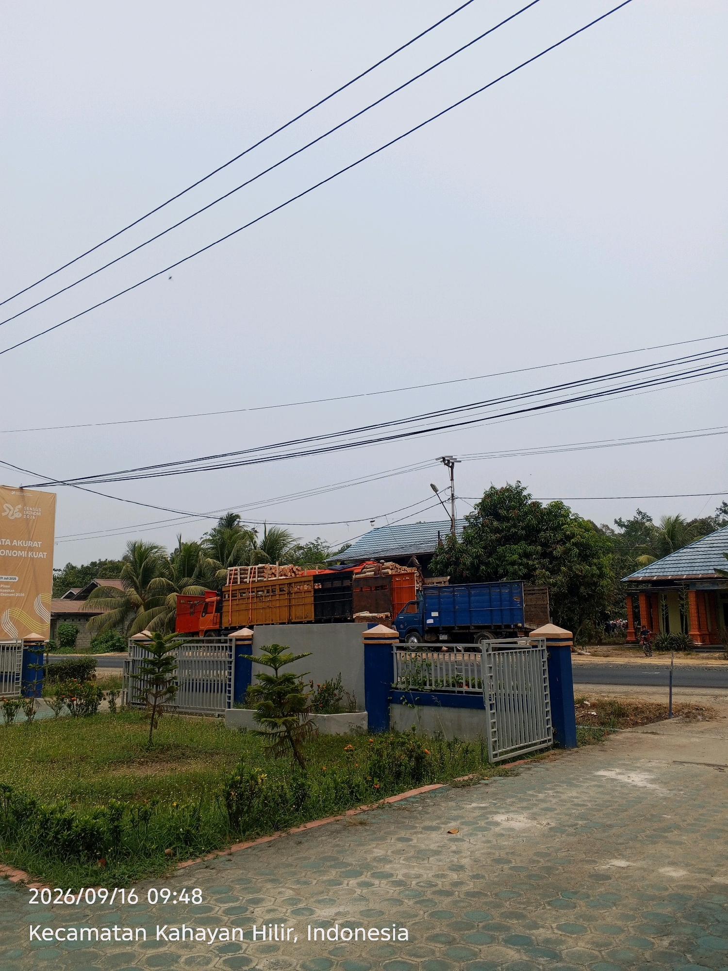 The image shows a street scene in the city of Mantaren II, Indonesia, on September 16, 2026, at 09:48 AM. The sky is overcast. Three power lines stretch across the upper portion of the frame. In the middle ground, two trucks are parked side-by-side on a paved area behind a low blue and beige wall topped with a metal gate. The truck on the left is orange and yellow and appears to be loaded with logs. The truck on the right is blue and has a wooden cargo bed. To the right of the blue truck is a building with a blue roof and orange brick accents. Behind the trucks and buildings, there are palm trees and other lush green vegetation. In the foreground, there is a patch of green grass with some small bushes and flowers, and a patterned paved walkway. A banner with text and graphics is visible on the left side of the frame, partially obscuring the orange truck. A person on a bicycle is visible in the far right background. The date and location are overlaid on the bottom left of the image.