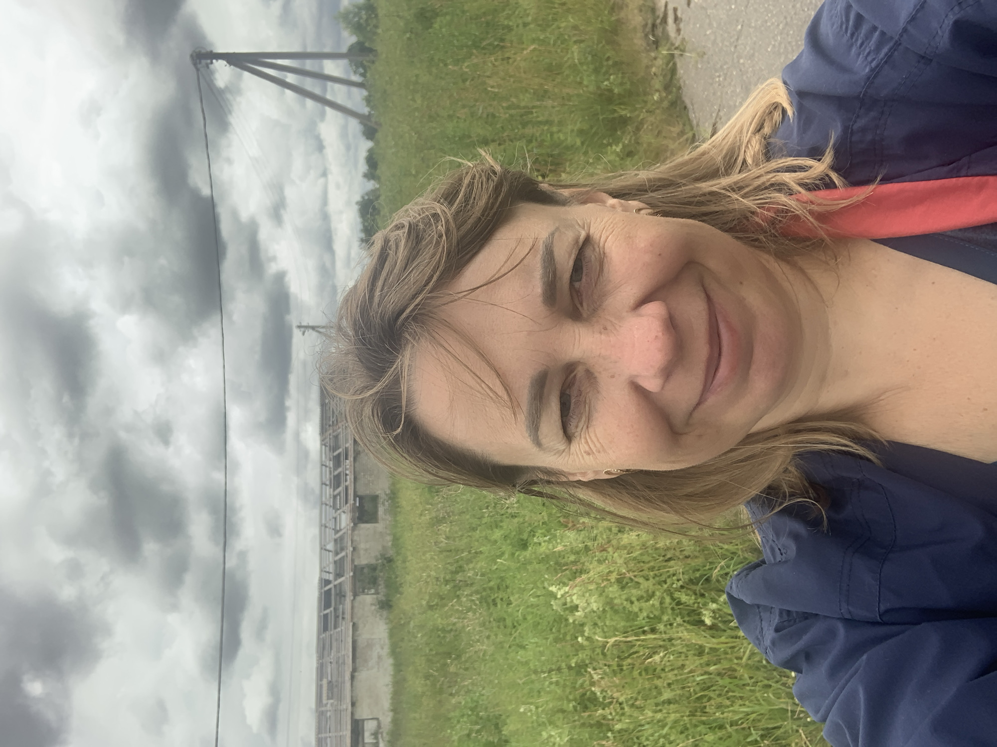 The image is a medium shot selfie taken outdoors in an open, rural setting under a cloudy sky. A middle-aged woman with light brown hair is the primary subject, smiling and looking directly at the camera. She is wearing a dark blue jacket with a red scarf or garment visible underneath. In the background, there is a large, dilapidated building with a damaged roof, suggesting it is abandoned or under construction. This building is situated in a field of tall, green grass and weeds. Electrical poles and wires are visible in the middle ground, and the ground in the foreground appears to be a gravel or dirt path. The overall atmosphere is overcast and slightly bleak, consistent with a cloudy day. The location is identified as Vidzavski Sel'ski Savet, Belarus.