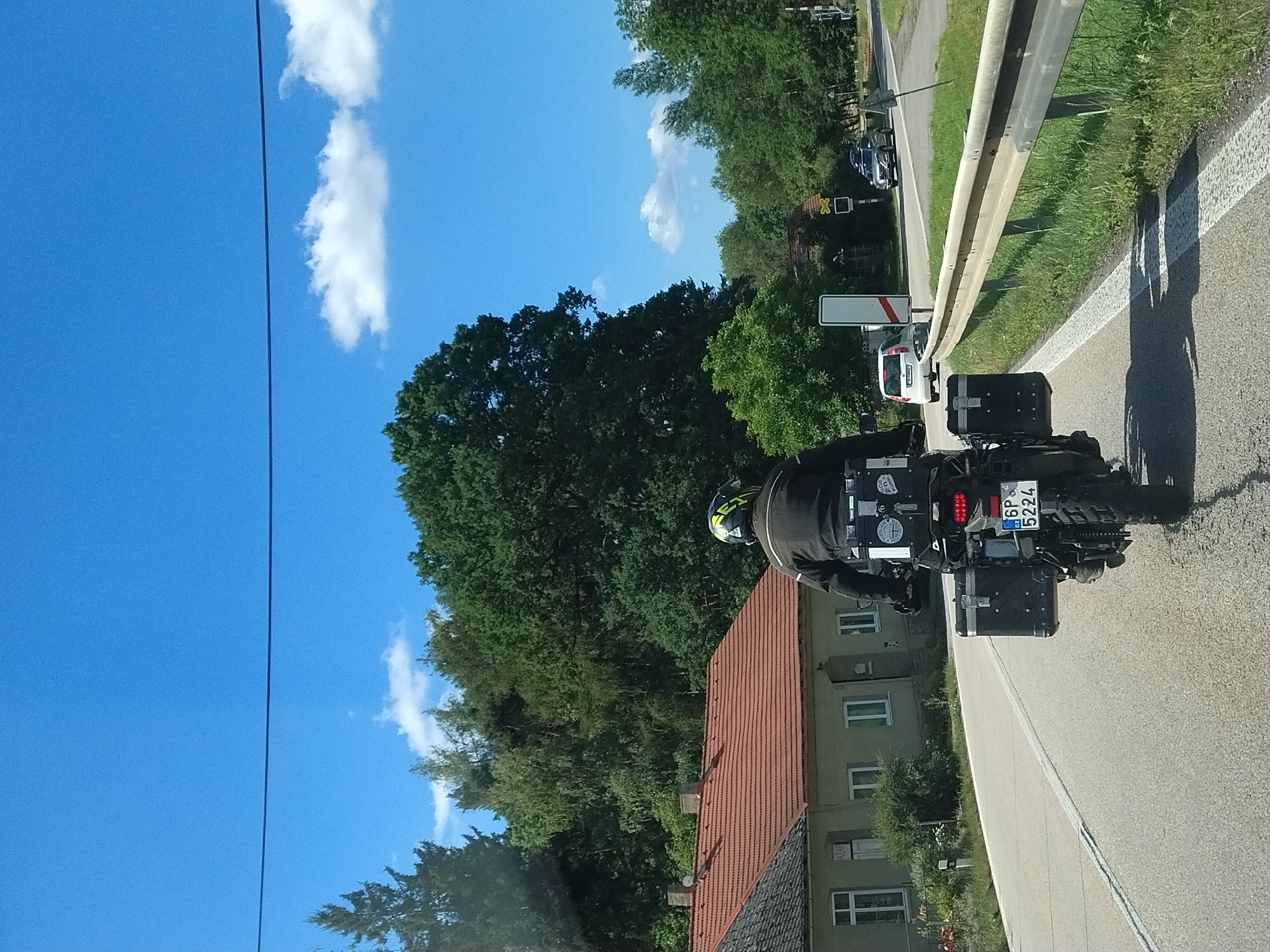 The image shows a rear view of a motorcyclist on a paved road in Strunkovice nad Volyňkou, Czechia. The motorcycle is equipped with two large black panniers on either side and a top box. The license plate reads "6P 5224". The motorcyclist is wearing a black jacket and a yellow and black helmet. To the left of the road, there is a light-colored building with a red tiled roof and a grey roof in the foreground. A wooden fence and grass are visible to the right of the road, followed by a metal guardrail. Trees and a clear blue sky with a few white clouds form the background. In the distance, another vehicle, a white car, is visible on the road, as well as a silver SUV. There are also road signs visible, including a railway crossing sign. The scene appears to be a sunny day.