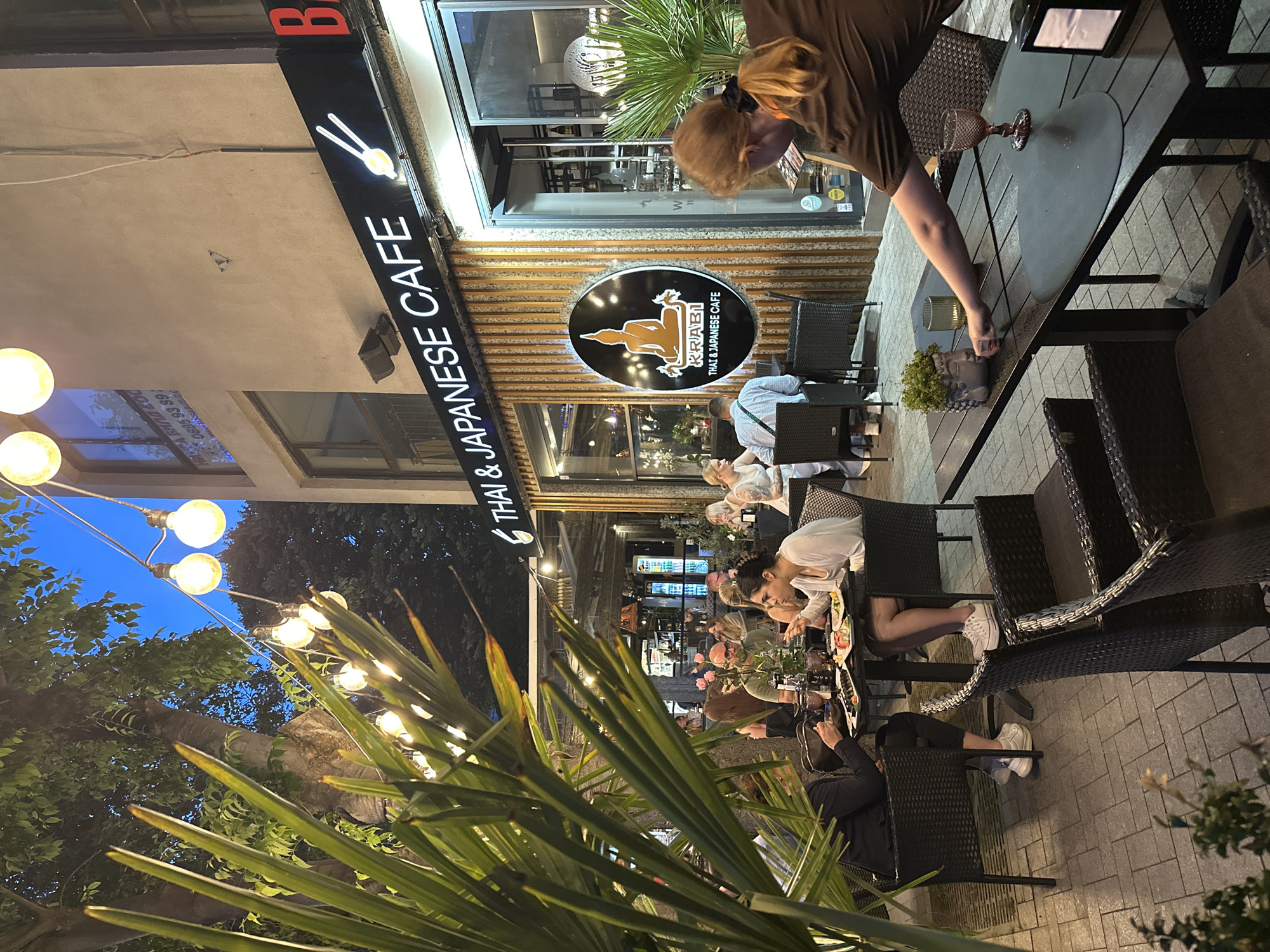 The image depicts an outdoor dining area of a Thai and Japanese cafe, identified as Krabi Thai & Japanese Cafe, located in Varna, Bulgaria. The scene is set at dusk or nighttime, illuminated by string lights and the interior lighting of the establishment. In the foreground, a waiter with blonde hair, wearing a brown t-shirt, is seen reaching across a dark wooden table, possibly attending to a guest or preparing the table. The table is set with placemats, a glass, and a small decorative item resembling a Buddha head with greenery. In the background, several patrons are seated at tables, engaged in dining and conversation. The facade of the cafe features a prominent sign with the restaurant's name and logo, a stylized Buddha figure. The outdoor seating area is furnished with dark wicker chairs and tables, and the ground is paved with gray stone tiles. Lush green foliage is visible in the left foreground, framing the scene. The overall atmosphere suggests a lively, urban dining experience.