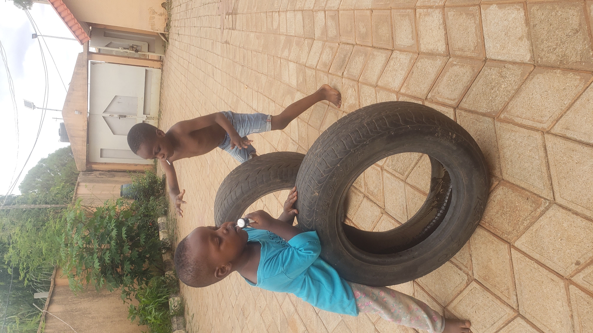 Two young Black children are playing with two large tires on a paved outdoor area in Sabon Tasha, Nigeria. The child on the left, a boy, is wearing a teal long-sleeved shirt and patterned leggings, and is leaning on one of the tires while holding a small white object to his mouth. The child on the right, also a boy, is shirtless and wearing denim shorts, and is reaching out a hand towards the other child. The tires are black and appear worn. The paving stones are tan and arranged in a geometric pattern. In the background, there is a wall, a large metal gate, and some vegetation. The sky is overcast.
