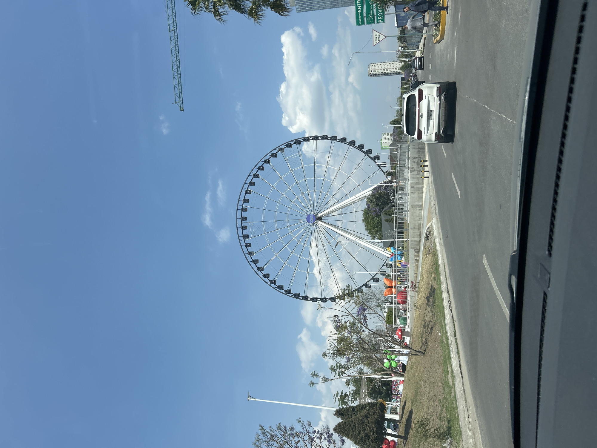 A sideways view from within a vehicle shows a large Ferris wheel dominating the skyline against a backdrop of a partly cloudy, blue sky. A white vehicle travels on a road alongside the Ferris wheel. Various signs, buildings, trees, and a green crane are visible in the background, with decorative structures near the base of the Ferris wheel.