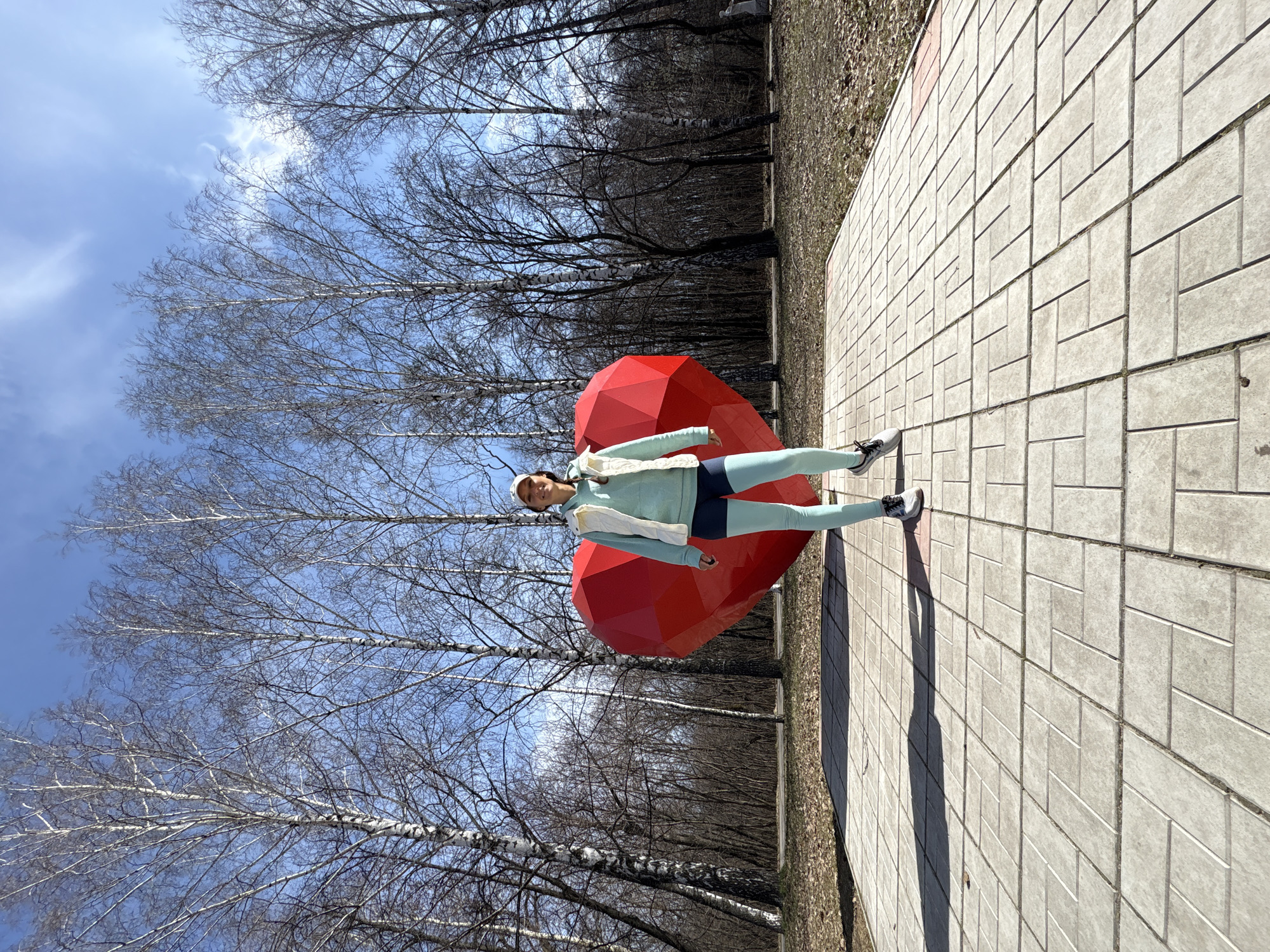 A full-body shot captures a woman standing next to a large, prominent red polygonal heart sculpture in an outdoor setting in Ulyanovsk, Russia. The woman, who appears to be in her 20s or 30s, is wearing light blue athletic leggings and a matching long-sleeved top or jacket, layered with a white quilted vest and a white baseball cap. She is also wearing athletic shoes. She stands with her left leg slightly forward and arms relaxed at her sides, looking towards the camera with a faint smile.
The background consists of numerous tall, bare deciduous trees, likely birches given the visible light bark on some branches, suggesting the season is late winter or early spring before the foliage has returned. The sky above is clear and blue with a few scattered, wispy white clouds. The ground on the right side of the image is covered in grey rectangular paving stones, on which the long shadows of the woman and the heart sculpture are cast, indicating bright sunlight. To the left of the paved area, there is dry ground covered with sparse grass and fallen leaves.
The scene depicts a sunny daytime outing in a park or public area. No visible text is present in the image.