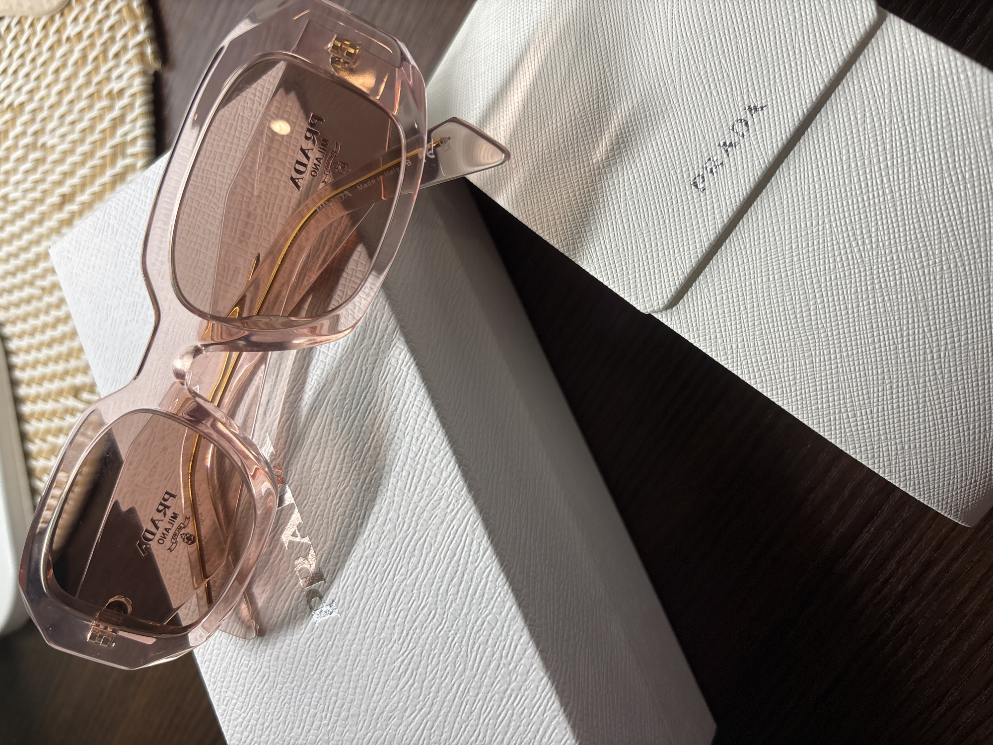 The image shows a pair of pink Prada sunglasses resting on a white textured box, with a white Prada envelope behind it. The background includes a dark wooden surface and a woven material. The image is well-lit. There is no indication of the location being Ungasan, Indonesia.