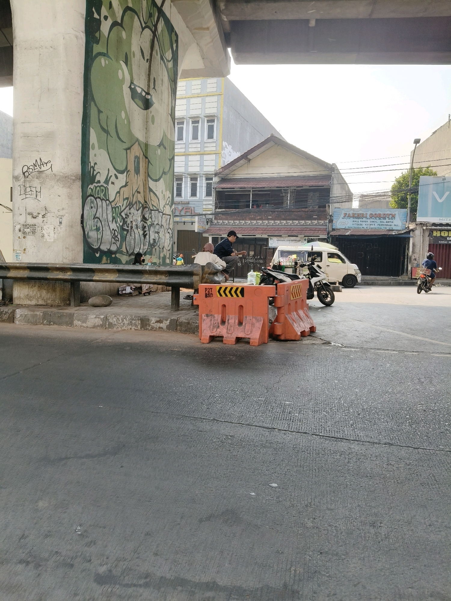 This image depicts a street scene in South Tangerang, Indonesia, under an elevated structure adorned with graffiti. A large column on the left features a green mural of a smiling tree with bold graffiti underneath. A metal guardrail runs horizontally across the mid-ground. On the sidewalk behind the guardrail, several individuals are present. Two people, one wearing a headscarf and another a light-colored shirt, are seated near the guardrail, appearing to rest or converse. Another person is partially visible to their left. In front of them are bags and possibly personal belongings.
The street itself is paved and appears to be a two-lane road. Orange plastic traffic barriers are positioned on the right side of the sidewalk, suggesting ongoing construction or a temporary road diversion. A white van is parked on the street, and a black motorcycle is positioned beside it. Further down the street, a person is riding a motorcycle.
Buildings line the street, including a multi-story building with a yellow facade and white window frames in the background, and lower-rise structures with awnings and signage. The sky is bright, indicating daylight, and the overall atmosphere suggests a busy urban environment. The graffiti on the pillar and the presence of street vendors and parked vehicles are characteristic of an urban setting in Indonesia.