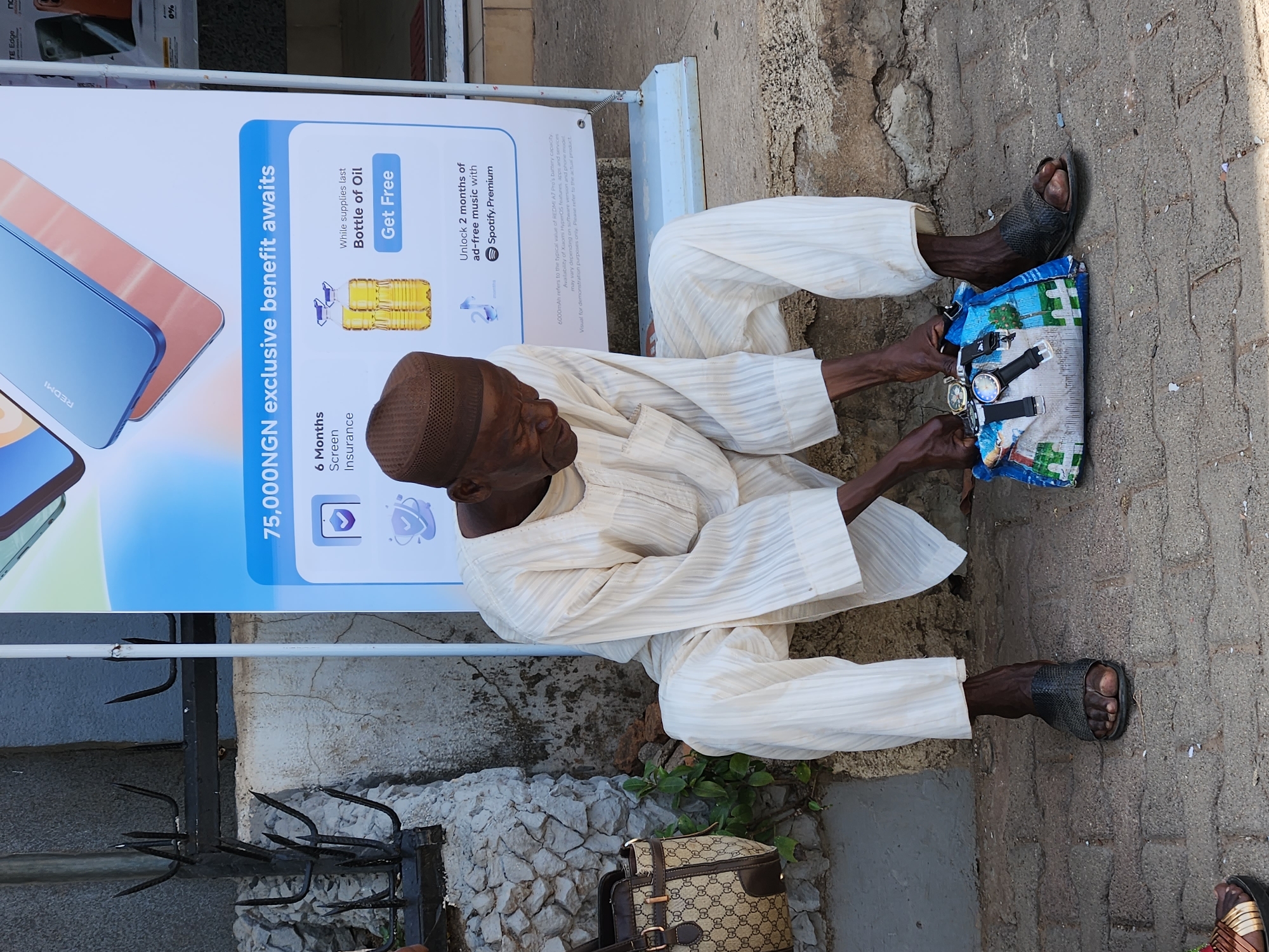 An elderly man is seated on the ground next to a promotional sign for a Redmi phone, on a city street of Unknown, Unknown. He wears a brown kufi cap, a white, long-sleeved striped traditional outfit, and dark-colored sandals. The man is holding a blue and white plastic bag displaying an assortment of watches. Near him is a tan Gucci handbag on the ground. The Redmi sign advertises a 75,000 NGN exclusive benefit, 6 months screen insurance, and a free bottle of oil, plus 2 months of ad-free music with Spotify Premium. The background shows a combination of a building wall, a black decorative metal fence, and a brick walkway.