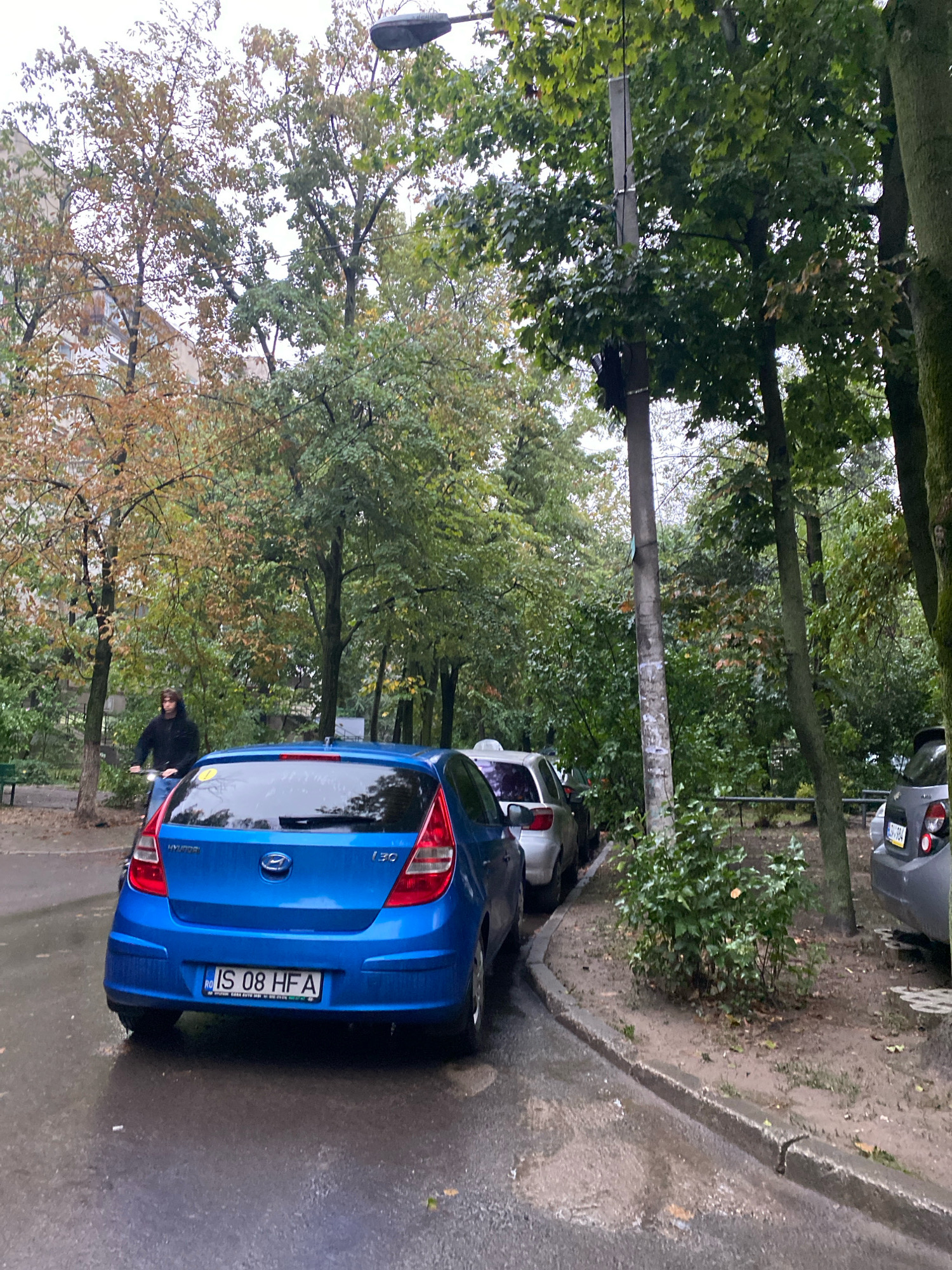 The image shows a street scene in Chișinău, Moldova. A blue Hyundai i30 car is parked on the right side of the street, facing left. The license plate reads "IS 08 HFA". To the left of the Hyundai, another car, a silver hatchback, is parked. In the background, several trees with autumn foliage are visible, along with a building. A person is seen riding a bicycle on the left side of the frame, wearing a black hoodie. The street is wet, suggesting it might be raining or has recently rained. A lamppost is positioned near the center of the image, alongside a utility pole. The overall environment appears to be a residential area with parked cars and green spaces.