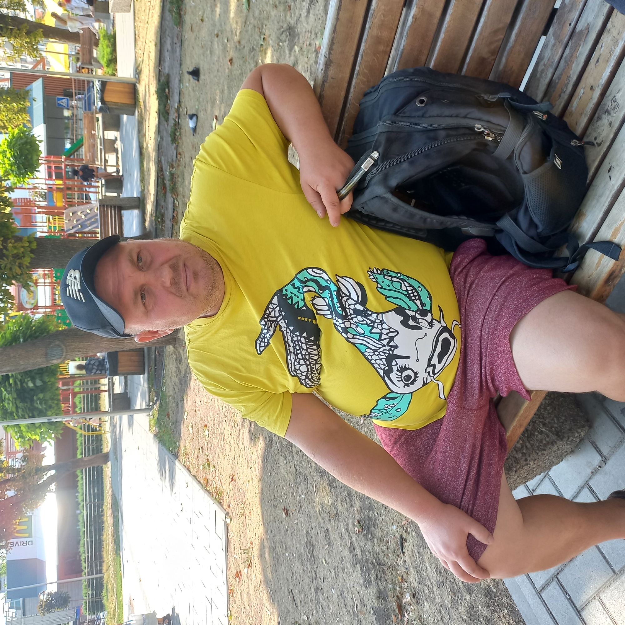 A man wearing a yellow t-shirt with a graphic print and a black baseball cap is sitting on a wooden bench in an outdoor park. He is holding a black backpack on his right side. The park appears to be in Kropyvnytskyi, Ukraine, given the context of other images provided from the same source. In the background, there is a playground with colorful equipment, trees, and other people. The ground is a mix of paved walkways and sandy/grassy areas. The lighting suggests it is daytime.