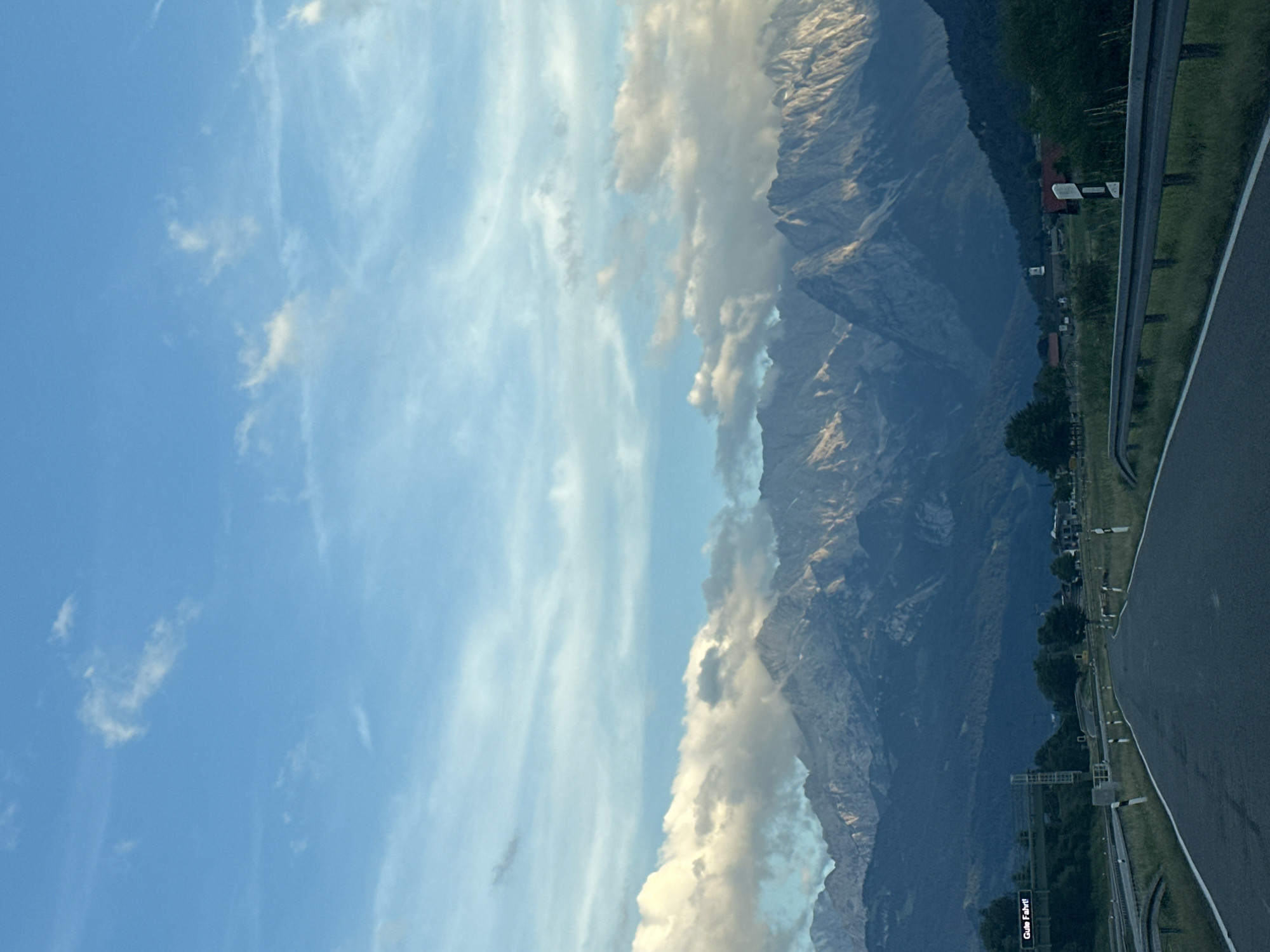 The image depicts a scenic view of the Alps in the vicinity of Farchant, Germany. A paved road curves towards a majestic mountain range partially obscured by clouds. The sky is a clear blue, with wispy clouds scattered across it. The mountains are rugged and imposing, with their peaks catching the light.
In the foreground, the road is bordered by guardrails and grassy verges. To the left of the road, an overhead sign displays the German phrase "Gute Fahrt!", which translates to "Have a good trip!". The environment appears to be a rural or semi-rural area, with some scattered buildings visible in the distance. The overall impression is one of a serene and picturesque landscape, ideal for a road trip.