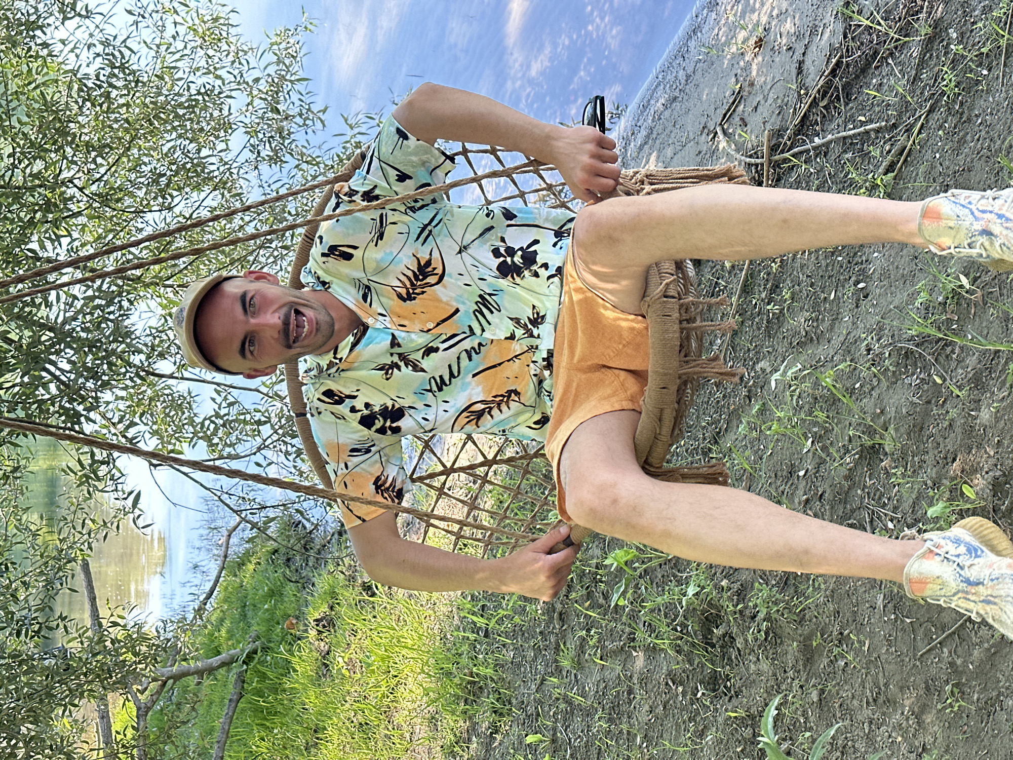 A man in a floral shirt, orange shorts, and sneakers is seated in a rope chair swing near a body of water, possibly in Vranjina, Montenegro. He is smiling and holding the sides of the swing. The area surrounding the swing is grassy with trees and foliage. The water reflects the sky.
