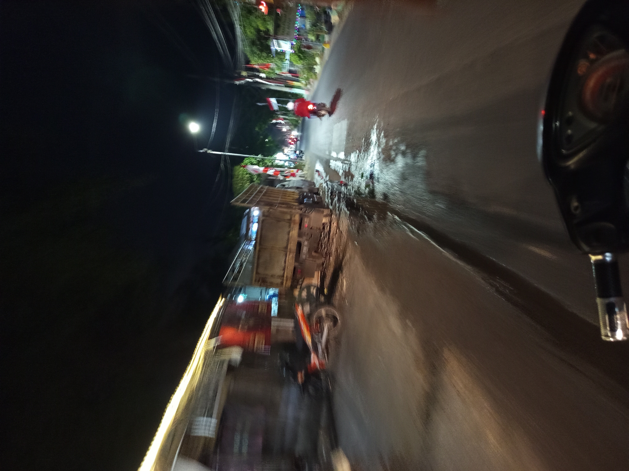 The image depicts a street scene at night in Punggul, Indonesia, likely captured from the perspective of someone riding a motorcycle. On the left side of the street, a parked truck is visible, with its bed appearing to be loaded with goods. Adjacent to the truck, a motorcycle is parked. The road surface is wet and reflects the streetlights and other illumination, suggesting it might have rained or there is standing water. Further down the street, another motorcycle rider wearing a red jacket is visible, moving away from the camera. The street is lined with trees and poles, some adorned with Indonesian flags. Buildings and shops are present along the roadside, illuminated by artificial lights. The overall impression is a busy street in a town or city at night.