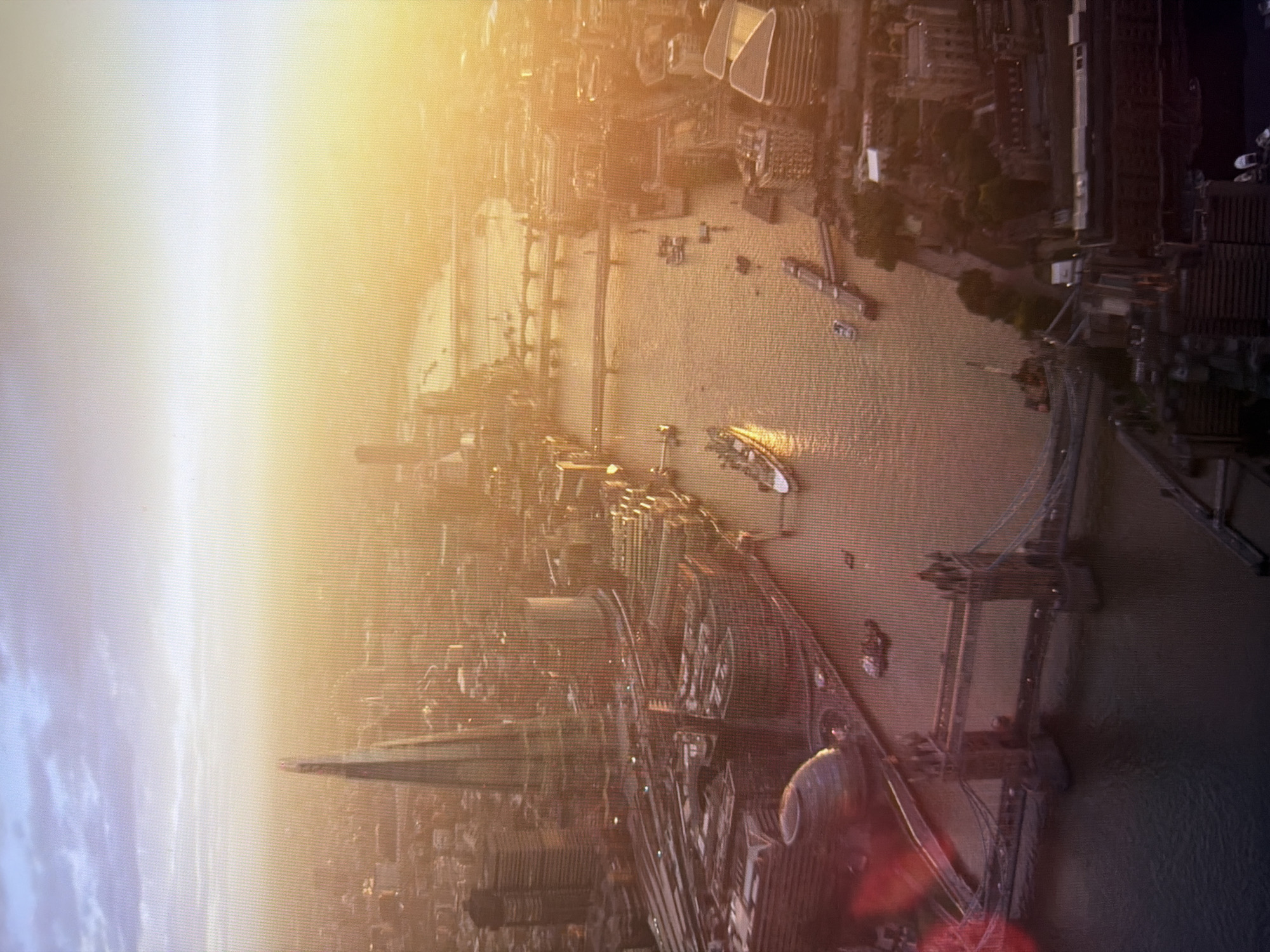 This is an aerial, high-angle view of a city at sunset, bathed in a warm, golden light. The dominant feature in the foreground is the Tower Bridge, a historic suspension bridge with two Gothic towers. The River Thames flows beneath it, reflecting the warm hues of the sky. Further upstream, other bridges cross the river, and several boats and a large ship are visible on the water. In the mid-ground, the iconic skyscraper "The Shard" pierces the skyline, its distinctive glass pyramid shape illuminated by the setting sun. Surrounding these landmarks is a dense urban landscape of buildings of varying heights and architectural styles. The sky transitions from a pale blue with wispy clouds near the top of the image to a bright, hazy orange and yellow as it meets the horizon.
The image does not depict Kyiv, Ukraine. The landmarks visible are consistent with London, United Kingdom, specifically The Shard and Tower Bridge.