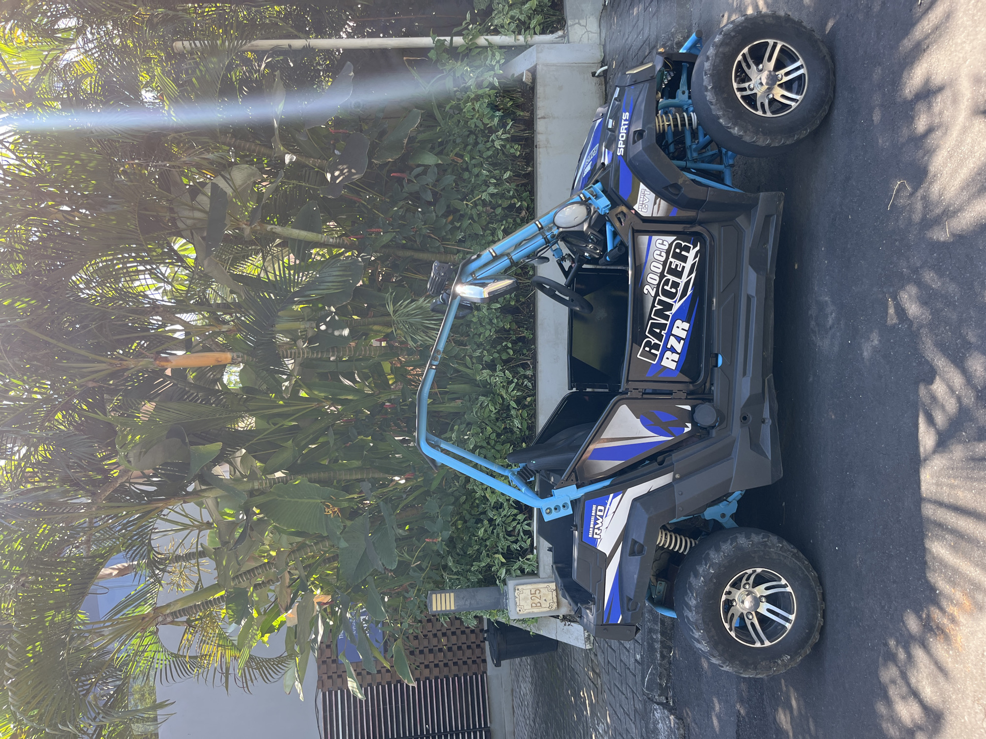 A side view of a blue and black RZR 200cc off-road vehicle is parked on an asphalt road in Kuta Selatan, Indonesia. The vehicle has a roll cage and a driver's seat. Behind the vehicle is a wall with vertical louvers and a concrete planter filled with lush green tropical plants, including palm trees. A black trash can is visible to the left of the vehicle. The sun is shining brightly, casting shadows on the road and illuminating the scene. The sky is not visible in the image.