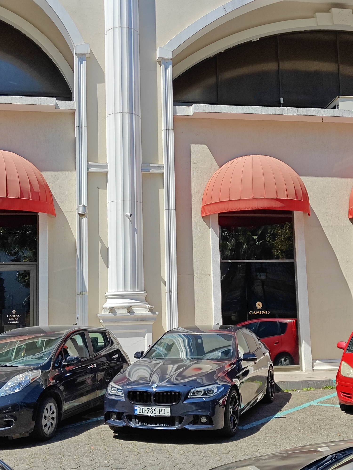 The image captures a street scene in Batumi, Georgia, likely in front of a casino. The building has an off-white facade with architectural details including arched windows, red awnings over doorways, and prominent white columns. Several cars are parked in front of the building, including a black Nissan Leaf, a dark blue BMW with license plate "DD-786-PD" (likely a Georgian license plate), and parts of two red cars are visible. The weather appears sunny, casting shadows on the building and street. The overall scene suggests a commercial or leisure area in the city.