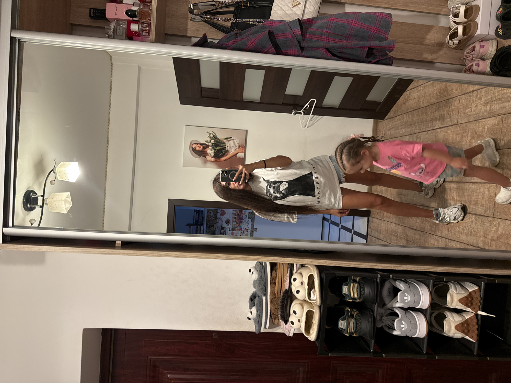 In a residential interior, a young woman stands in front of a full-length mirror, taking a selfie with her phone. She is wearing a white t-shirt with a dog print and denim shorts. Next to her, a young child in a pink t-shirt and denim shorts walks away from the mirror, viewed from the side. The mirror reflects a tidy room with a wooden door on the left, a shoe rack filled with various footwear, and shelves displaying perfumes and bags on the right. A framed picture of a woman holding tulips hangs on the wall behind the young woman. The floor is made of light-colored wood planks. The lighting suggests it is daytime, with overhead lights also visible in the reflection. The overall impression is of a personal dressing or hallway area within a home.
