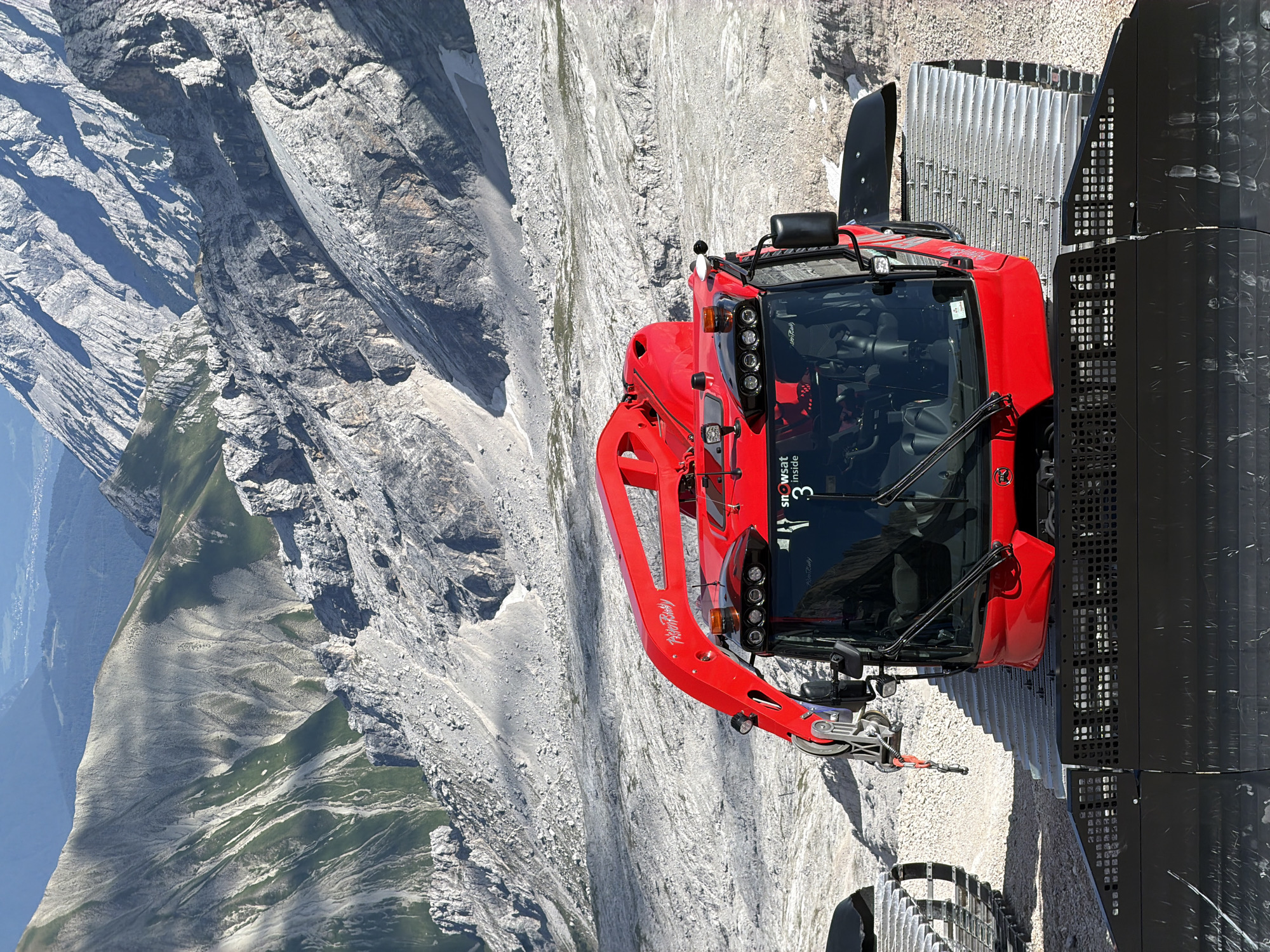A bright red snow grooming vehicle, a PistenBully 300, is parked on a gravelly surface in a mountainous area. The vehicle has large tracks, a driver's cabin with multiple lights, and a mechanical arm extending from the top. The driver's cabin has a windshield with wipers and side mirrors. On the windshield, there is a sticker that reads "SNOWsat inside 3". The vehicle is positioned in front of a steep, rocky mountain slope with patches of green vegetation. The background shows more rugged mountain ranges under a clear blue sky. The overall environment suggests a high-altitude location, likely a ski resort or alpine region. While the image appears to be taken in Wildermieming, Austria, there are no people or specific city landmarks visible in the frame.