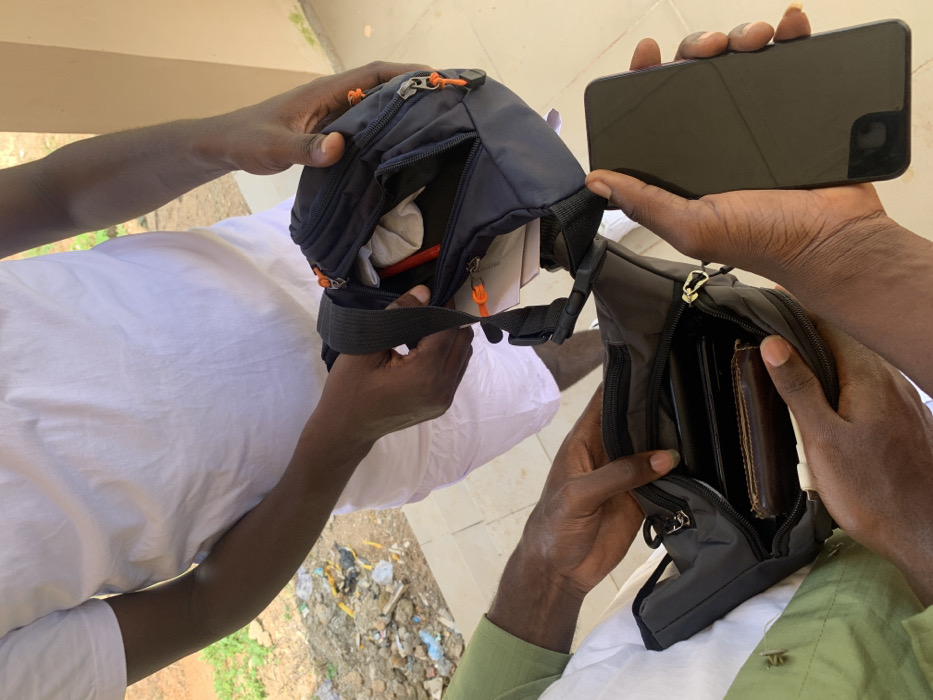 The image captures three individuals in an outdoor setting in Kubi A, Nigeria. The central figure, wearing a white t-shirt and white trousers, holds a black fanny pack with orange zipper pulls open, revealing a crumpled white item and a red cylindrical object within. A second individual, on the lower left and wearing a green shirt with white trousers, holds a similar black fanny pack with silver zipper pulls open, displaying a brown leather wallet and other dark objects inside. A third individual, on the lower right, holds a dark-colored smartphone with a blank, reflective screen, positioned towards the fanny packs. The ground is paved, and the background shows a dirt area with scattered refuse and some vegetation to the left, and a light-colored building wall with green discoloration to the right.