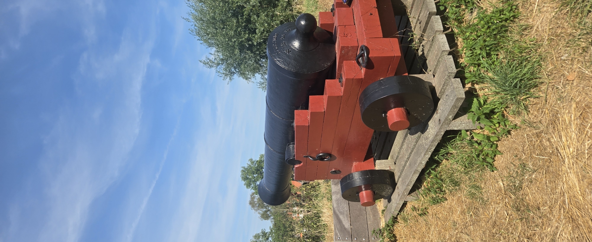 An antique black cannon is displayed outdoors on a red wooden carriage with black wheels. The cannon is positioned on a wooden platform, which is situated on a dry, grassy field. In the background, there are trees and a bright blue sky with scattered clouds. The scene appears to be a historical exhibit, possibly in the city of Brill, Netherlands, given the context. There are no people present in the image.