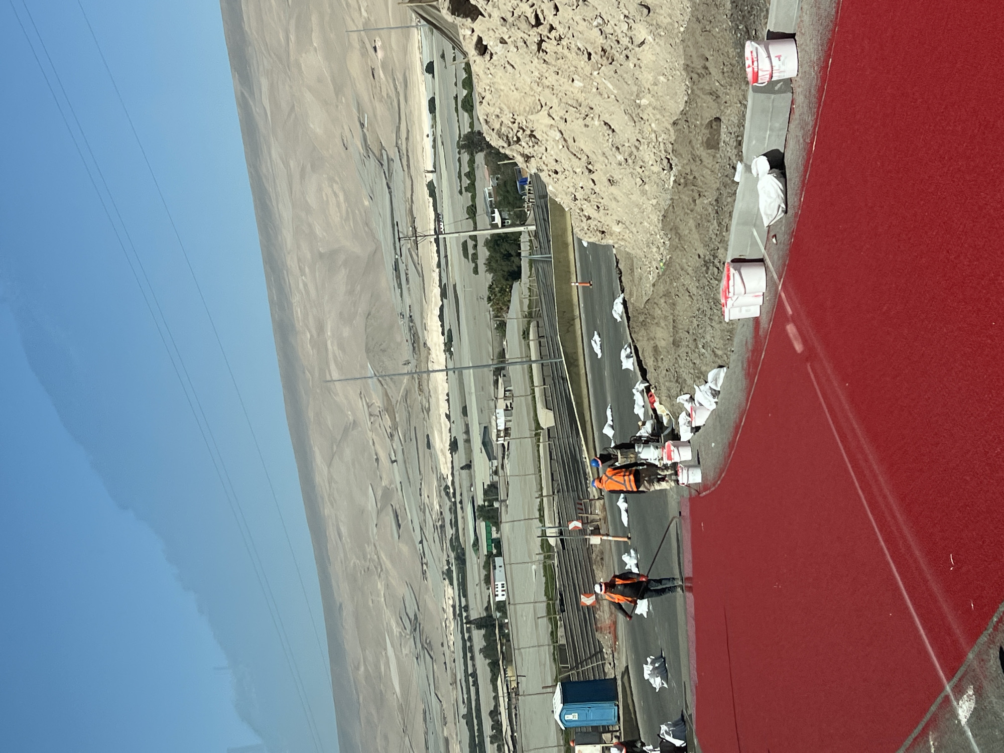The image depicts the ongoing repainting of a section of the Pan-American Highway in Arica, Chile. The roadway is being painted a bright red color. Two workers wearing orange safety vests are present; one is using a long-handled paint roller to apply the red paint to the road surface. Various white paint cans and white plastic bags, presumably used to protect the surrounding area from overspray, are scattered along the side of the road. A blue portable toilet is visible. The highway runs alongside a desert landscape on one side and a cliff or embankment on the other. The sky is clear and blue. Power lines are visible in the upper left corner of the image.