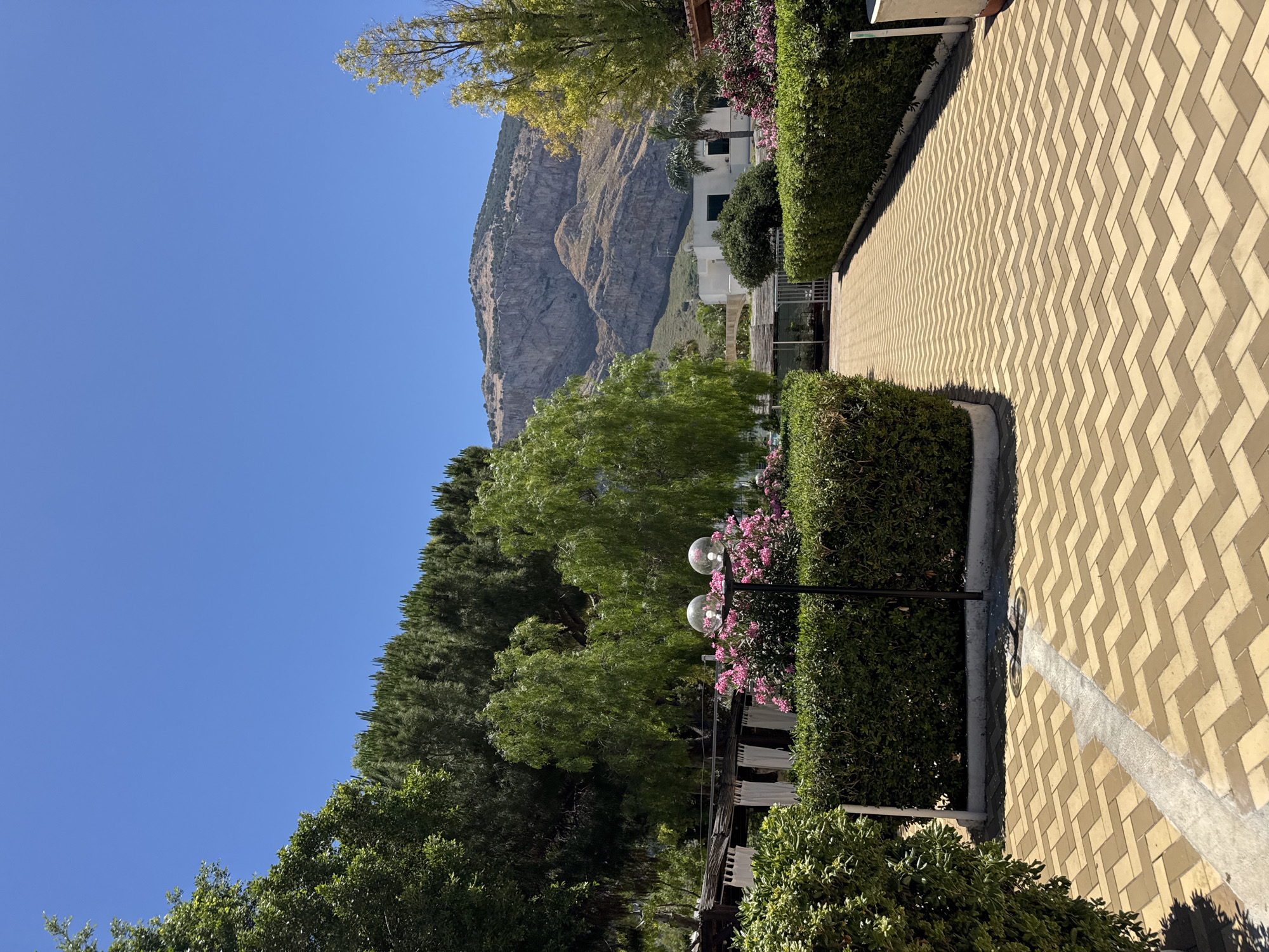 A bright, clear day is depicted in Isola delle Femmine, Italy, showcasing a well-maintained outdoor area. A path paved with beige and brown interlocking bricks, arranged in a chevron pattern, leads the viewer into the scene. This pathway is flanked by neatly trimmed green hedges. To the left, a portion of a dense green tree is visible, while to the right, the hedges continue, with pink flowers blooming in the background. A black lamppost with two spherical glass lights stands amidst the flowers. Further back, a white building with dark windows is nestled against a backdrop of a rugged, sun-drenched mountain range. The sky above is a vibrant, cloudless blue.