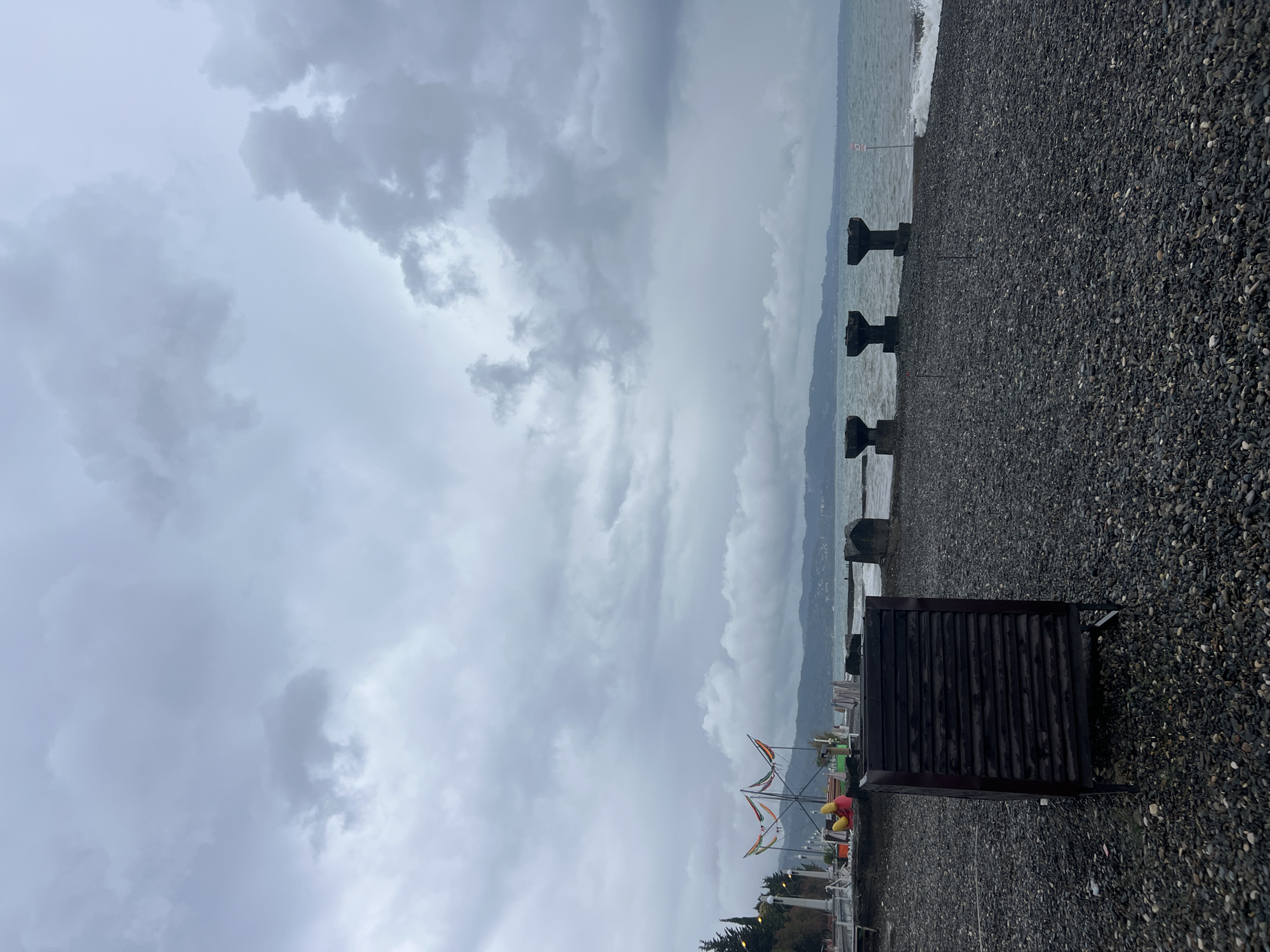 The image shows a dark, cloudy sky over a pebble beach in Sokhumi, Abkhazia. In the foreground, there is a dark wooden trash receptacle on metal legs. Beyond the receptacle, the beach is covered in dark pebbles. In the middle ground, the sea is visible with moderate waves. Several concrete pillars, likely part of a pier or breakwater, stand in the water. In the background, a distant coastline with mountains is shrouded in mist. To the left, a promenade with some lights and what appear to be colorful decorations is visible, along with some trees.