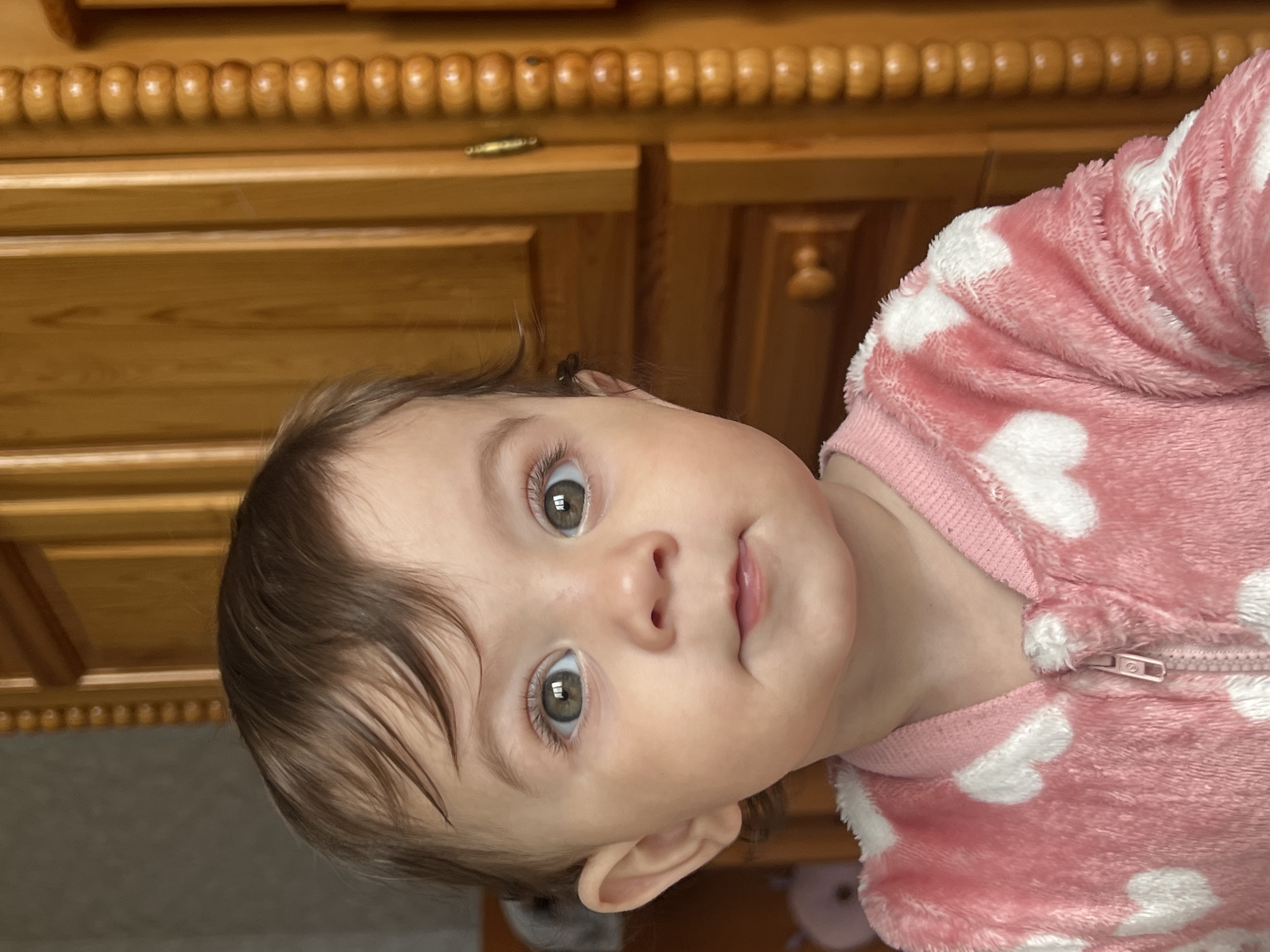 A close-up, tilted, eye-level shot features a young child, likely an infant, wearing a pink, plush garment decorated with white heart shapes. The garment appears to have a zipper closure. The child has fair skin, brown hair with bangs, and brown eyes. The child is looking directly at the camera with a neutral expression. In the background is a light brown wooden cabinet with decorative trim. The location is unknown, as there is no identifying information indicating Elena, Bulgaria, within the provided image.