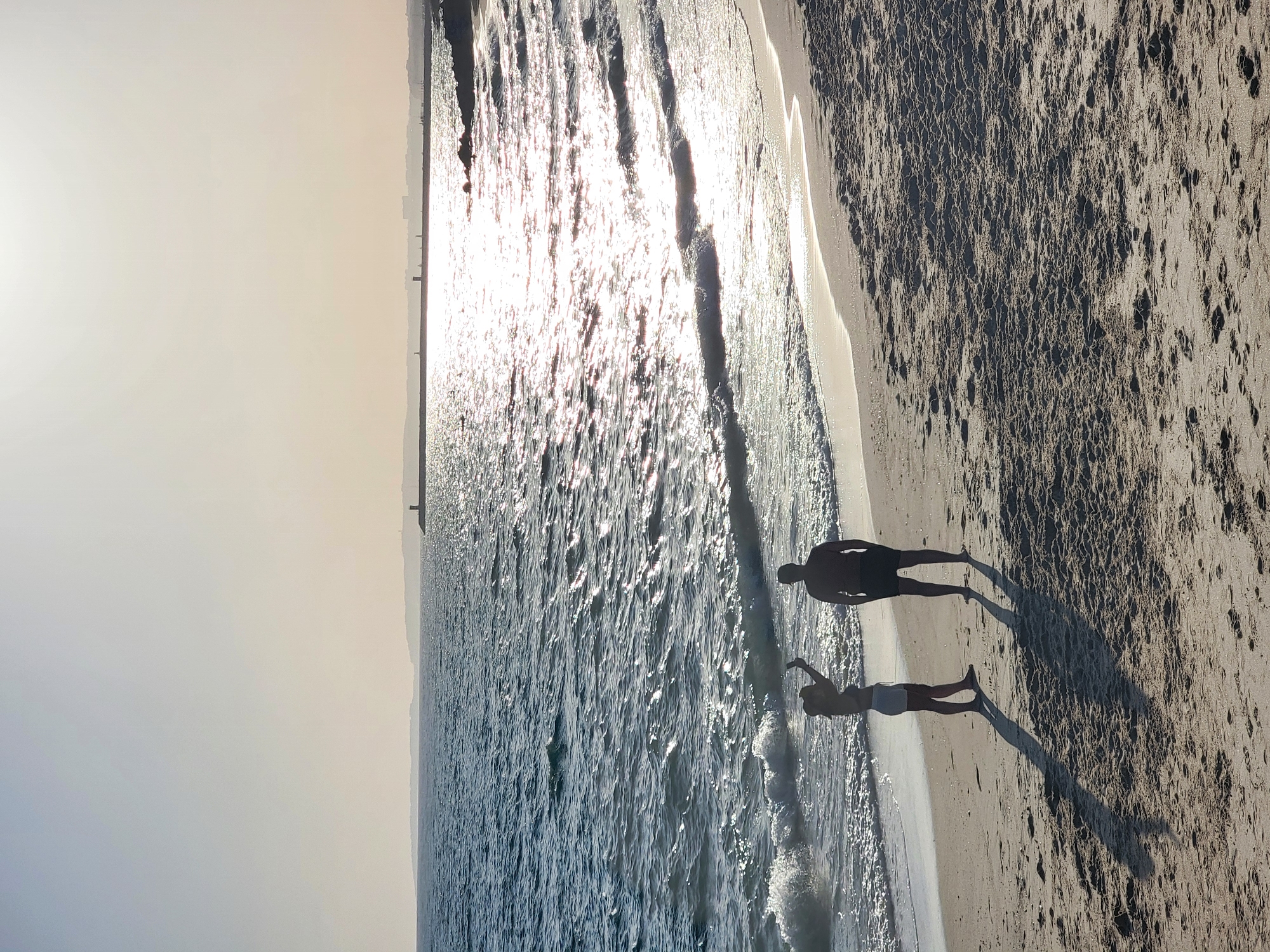 The image captures a serene beach scene in Quarteira, Portugal, likely during the late afternoon or early evening, given the low angle of the sun and the long shadows cast on the sand. The ocean, a deep blue, is dotted with shimmering reflections from the setting sun. Gentle waves lap at the sandy shore, leaving a wet, glistening edge.
In the foreground, two silhouetted figures stand on the beach. A person on the left, appearing to be taking a selfie with a phone held aloft, is dressed in light-colored shorts and a dark top. To their right stands another figure, a person with a more athletic build, wearing dark shorts and a dark t-shirt. Both figures are positioned with their backs to the camera, facing the ocean and the distant horizon.
The beach itself is composed of fine, light-brown sand, marked with the textured patterns of wind ripples and the occasional footprint. In the distance, a long, man-made breakwater or pier stretches across the water, punctuated by what appear to be small structures, possibly lighthouses or other navigational aids. Beyond the breakwater, a hazy outline of a town or coastline is visible against the bright sky. The overall atmosphere is peaceful and tranquil, conveying a sense of calm on a summer day.
