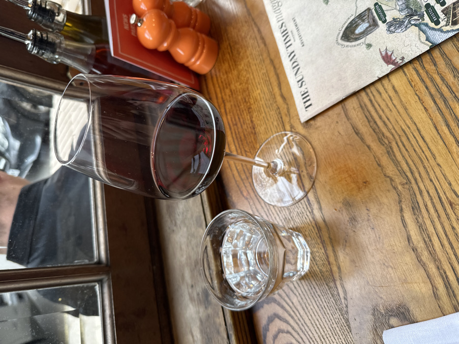 An indoor scene depicts a light brown wooden table, likely within a restaurant or pub in the City of London, United Kingdom. Centrally positioned on the table is a clear stemmed wine glass containing red wine. To its left, a clear, faceted tumbler holds water. In the upper right background, two clear glass bottles with pour spouts, one containing reddish liquid (likely vinegar) and the other yellowish (likely oil), stand next to two orange, conical salt and pepper shakers. A red menu holder is partially visible behind these condiment items. In the lower right, an open section of "THE SUNDAY TIMES magazine" lies flat, displaying an illustrated map with text including "GLOUCESTER" and "BRISTOL." A folded white napkin is partially visible in the lower left corner. A wooden-framed window in the background reflects the back of a person's head and shoulder, wearing a dark shirt, along with indistinct elements of an exterior street scene including a vehicle.