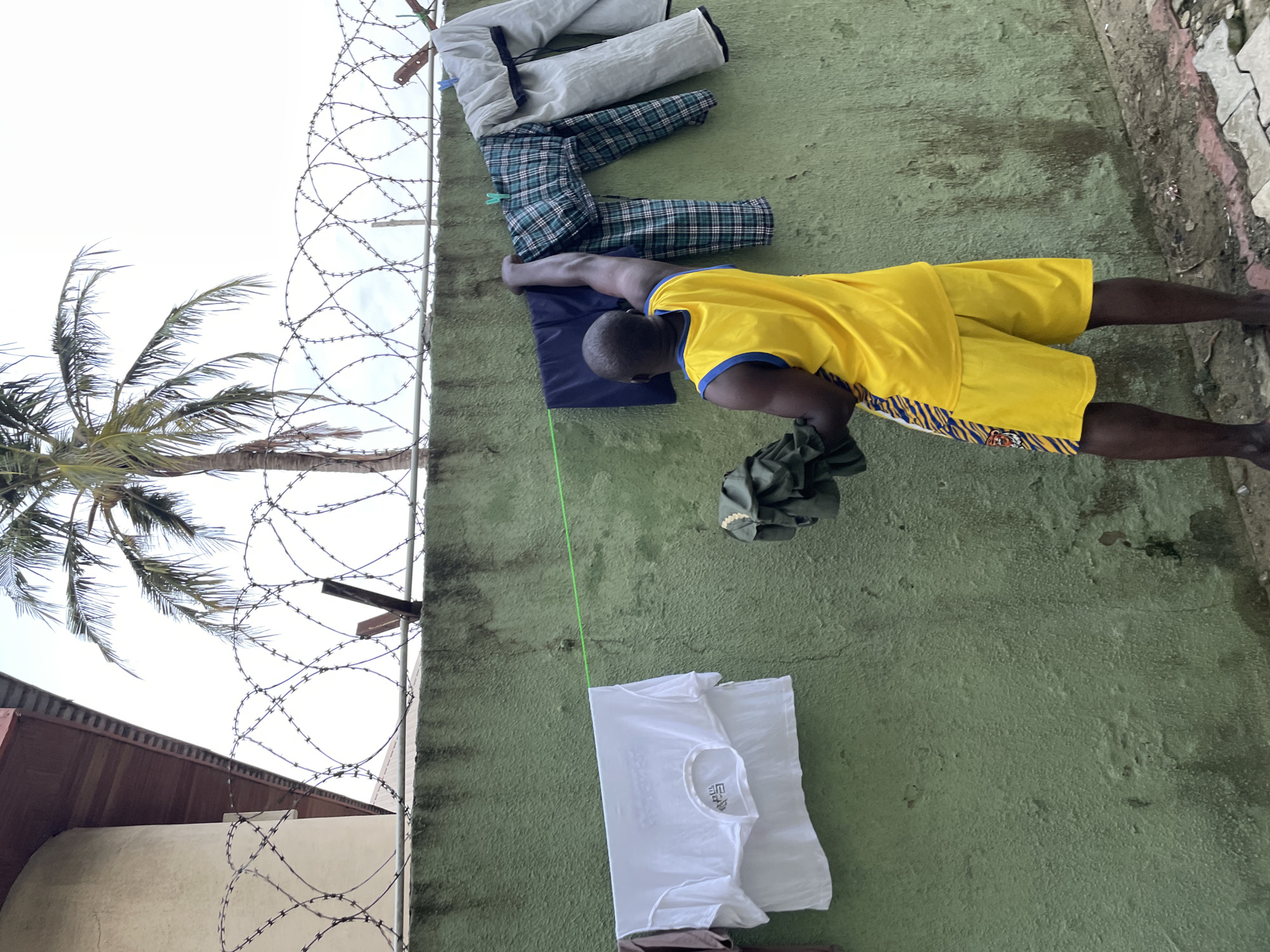 A Black man with dark skin and a muscular build is hanging laundry on a clothesline in Ado, Nigeria. He is wearing a bright yellow tank top with blue trim and yellow shorts with a tiger stripe pattern. He is holding a green garment in his left hand and reaching up with his right hand to adjust a navy blue garment on the clothesline. Several other items of clothing are hanging on the line, including white t-shirts, plaid pants, and grey pants. The clothesline is attached to a green, textured wall. Above the wall, there is a barbed wire fence, and behind that, a palm tree and a cloudy sky. The ground in front of the wall is a rough, paved surface with some debris. The scene appears to be an outdoor space, possibly a yard or alleyway, in a residential area of the city.