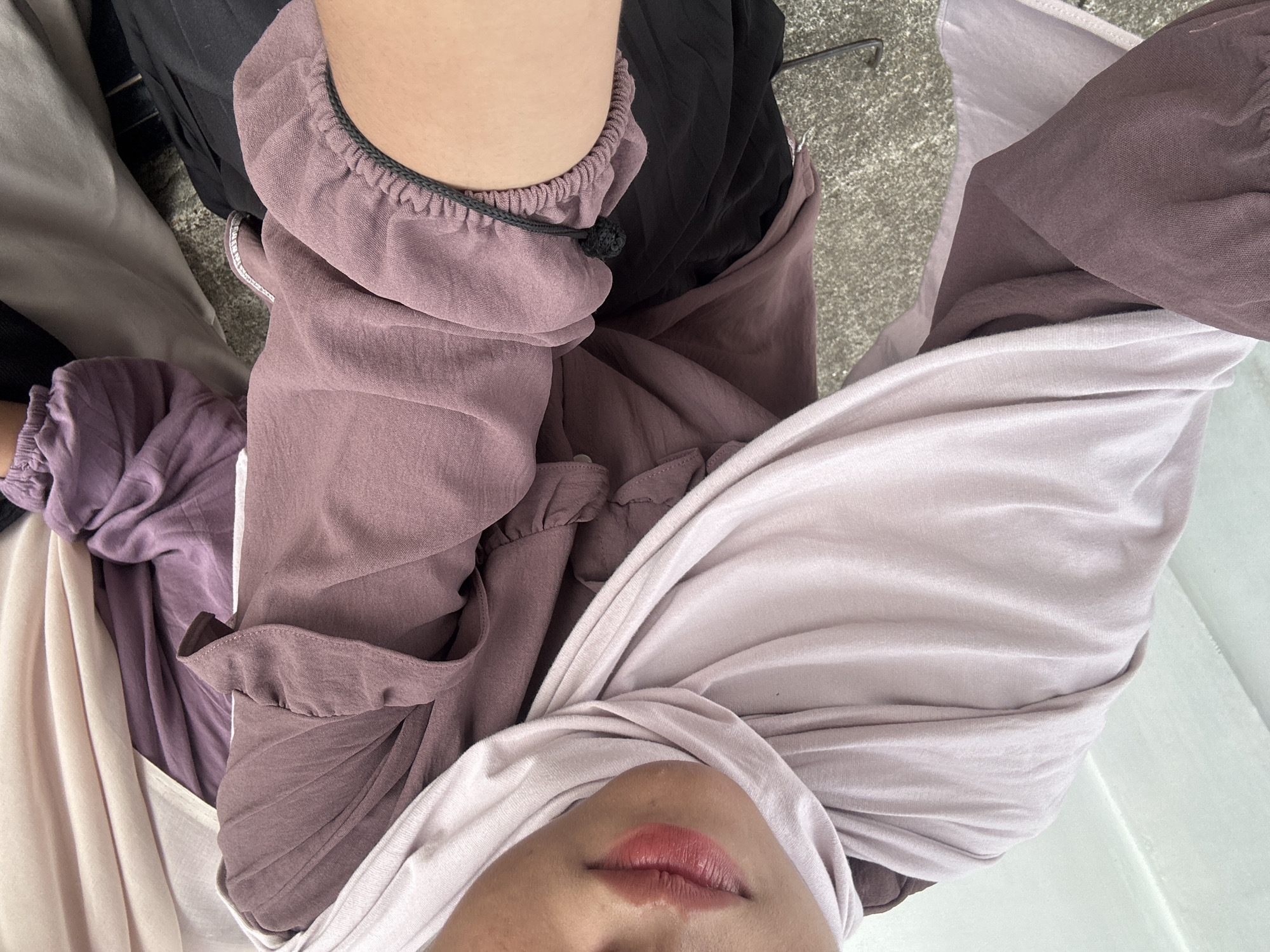 The image shows a close-up of a person wearing a light purple hijab and a dusty rose colored top with ruffles on the sleeves. The person also appears to be wearing a black skirt. The background consists of a grey, textured surface that resembles pavement or concrete. The lighting suggests an outdoor environment, possibly during the day. The scene is focused on the clothing and the person's face, specifically their lips which are painted with a glossy reddish-pink lipstick. The location is Palohlada, Indonesia.