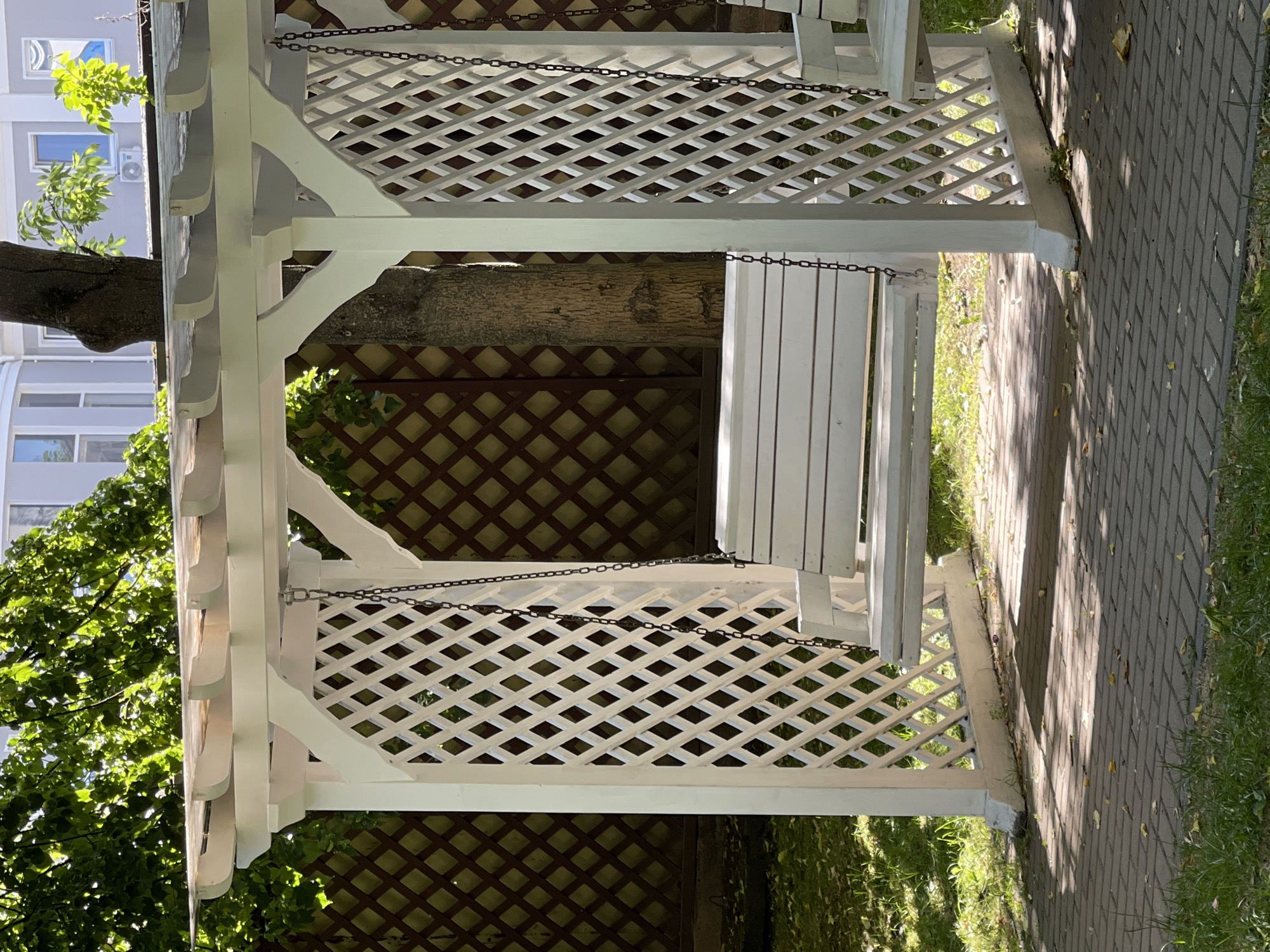 This media file depicts an outdoor scene featuring a white wooden gazebo with a swing. The gazebo has a latticework design on its sides and a slatted roof. A white swing, suspended by chains, is the central object within the structure. Sunlight filters through the leaves of a nearby tree, casting dappled shadows on a paved walkway and grassy areas surrounding the gazebo. The time of day appears to be daytime, with bright sunlight indicating fair weather. The presence of a building with windows in the background suggests an urban or suburban setting. The scene evokes a peaceful and tranquil atmosphere, likely in a park or residential garden. There are no people visible in the image, and no text is present. The location context is Zelenogradsk, Russia.