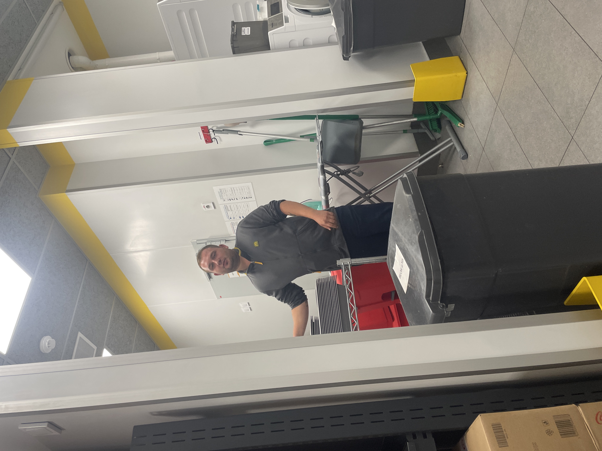 A man wearing a grey McDonald's jacket is standing in what appears to be a utility or laundry room. He is looking towards the camera with a neutral expression. To his left, a stack of grey plates is visible on a red shelf. In the background, cleaning supplies such as a mop and broom are leaning against a wall. There are also large black trash bins and white washing machines. The room is brightly lit with fluorescent ceiling lights and features yellow accent lines along the walls and ceiling. The overall environment suggests a commercial setting like a fast-food restaurant or a public facility. There is no discernible indication of the specific location within Boyarka, Ukraine, beyond the general appearance of an indoor utility space.