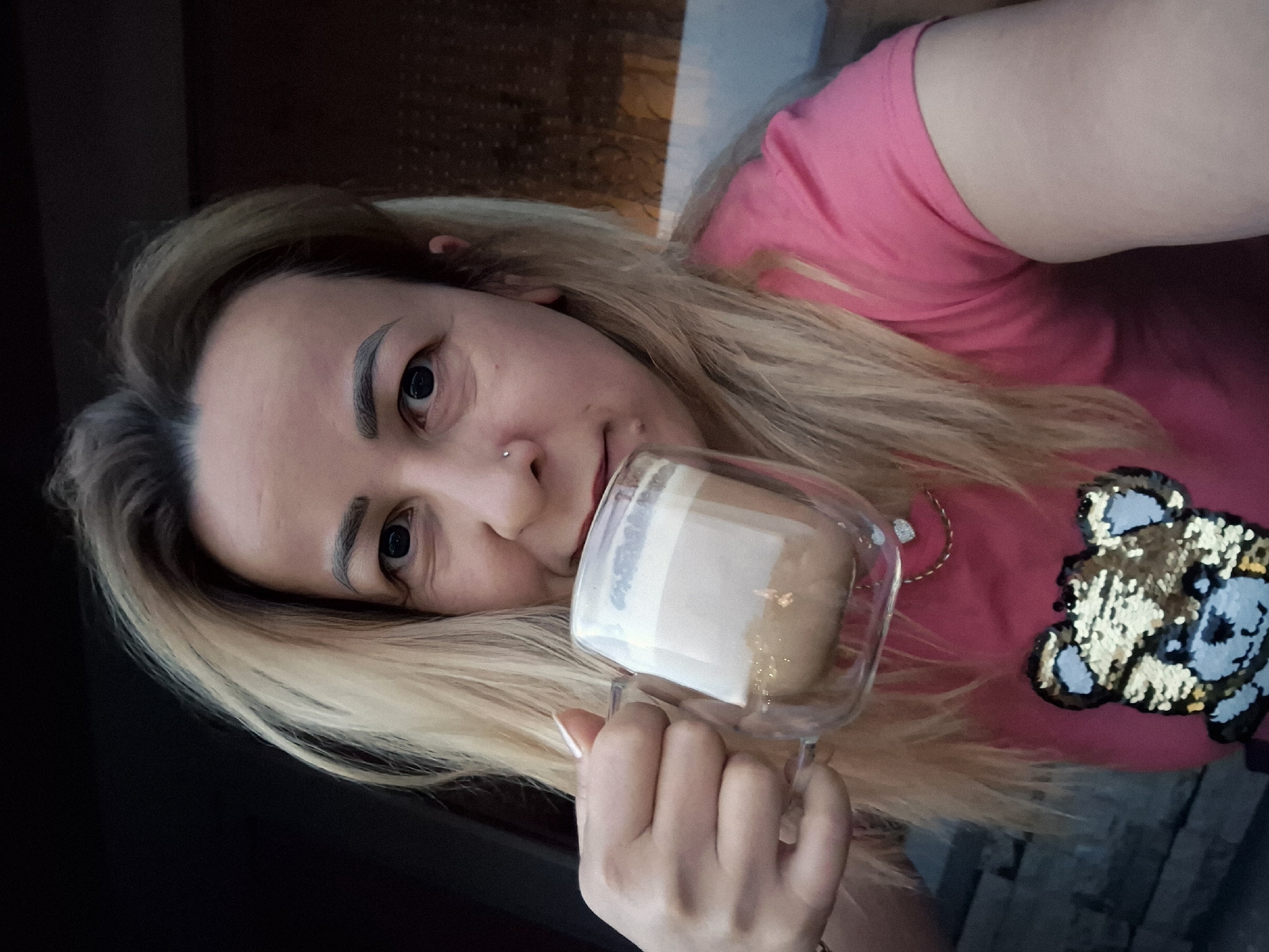 In a close-up, waist-up shot, a woman with blonde hair is captured from the chest up, holding a clear glass mug of coffee with both hands. She is wearing a bright pink t-shirt adorned with a sequined bear emblem. Her gaze is directed towards the camera, and she is in the process of taking a sip from the mug. The background is indistinct, suggesting an outdoor setting.
