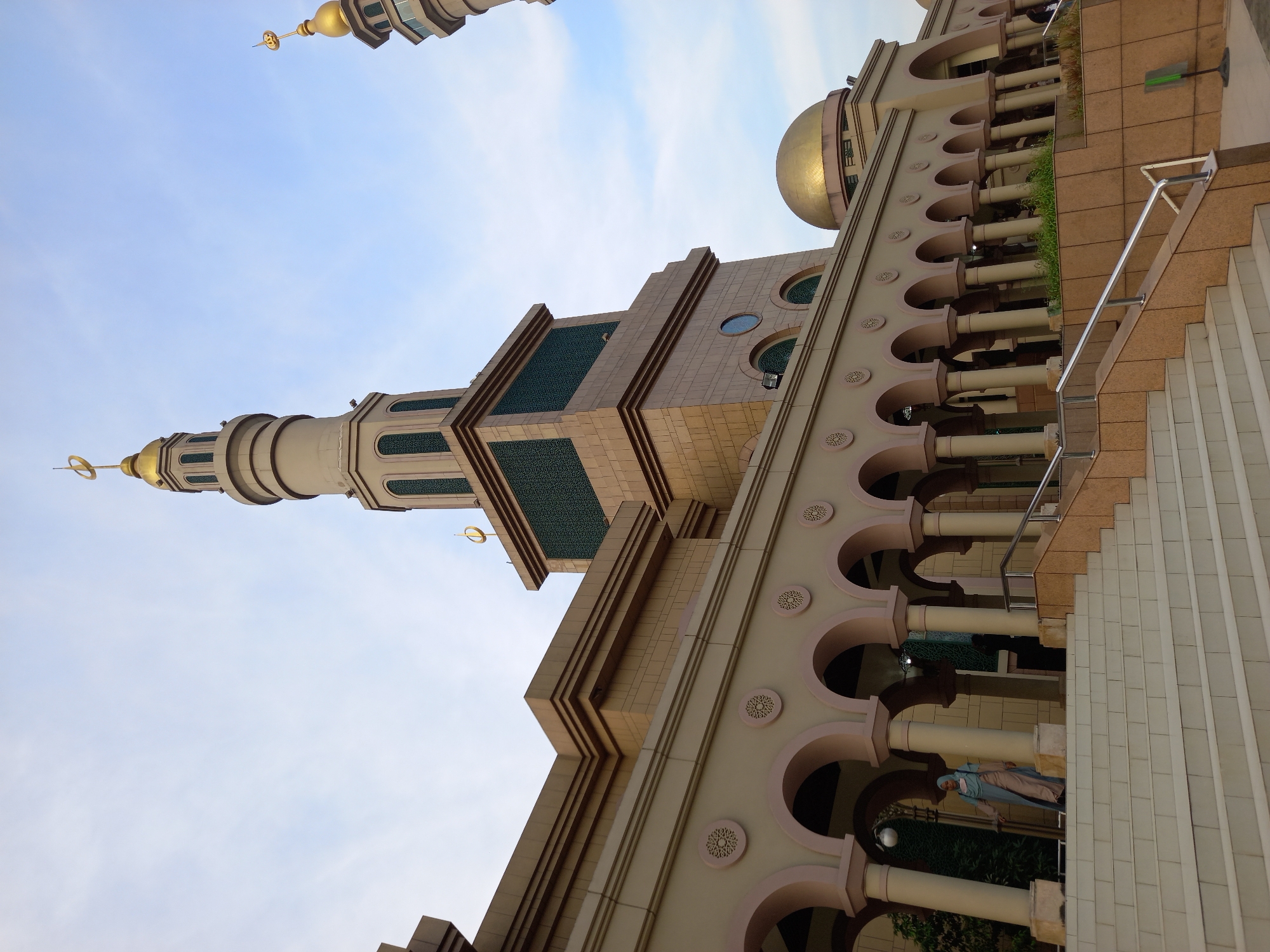 This is a low-angle, medium shot of the exterior of the Islamic Center Samarinda mosque in Samarinda, Indonesia. The building features a prominent, tall minaret with a gold dome and crescent moon at its apex, along with a smaller, gold-domed structure in the background. The mosque's facade is characterized by a colonnade of arched walkways with ornate circular designs above the arches. A wide, white staircase leads up to the mosque's entrance, and a woman wearing a hijab walks down the stairs on the left side of the image. The sky is partially cloudy.