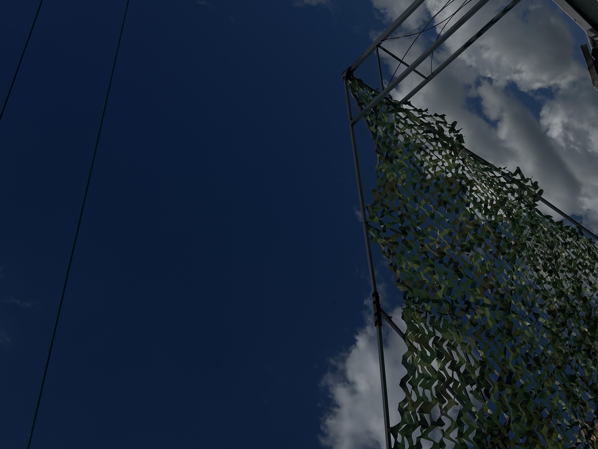 A camouflage net is stretched over a metal frame against a deep blue sky with scattered white clouds. The frame appears to be part of a structure, possibly a temporary shelter or a part of an outdoor installation. The scene is devoid of people or any discernible activity. There is no information available to pinpoint the location within the city of Shumen, Bulgaria.