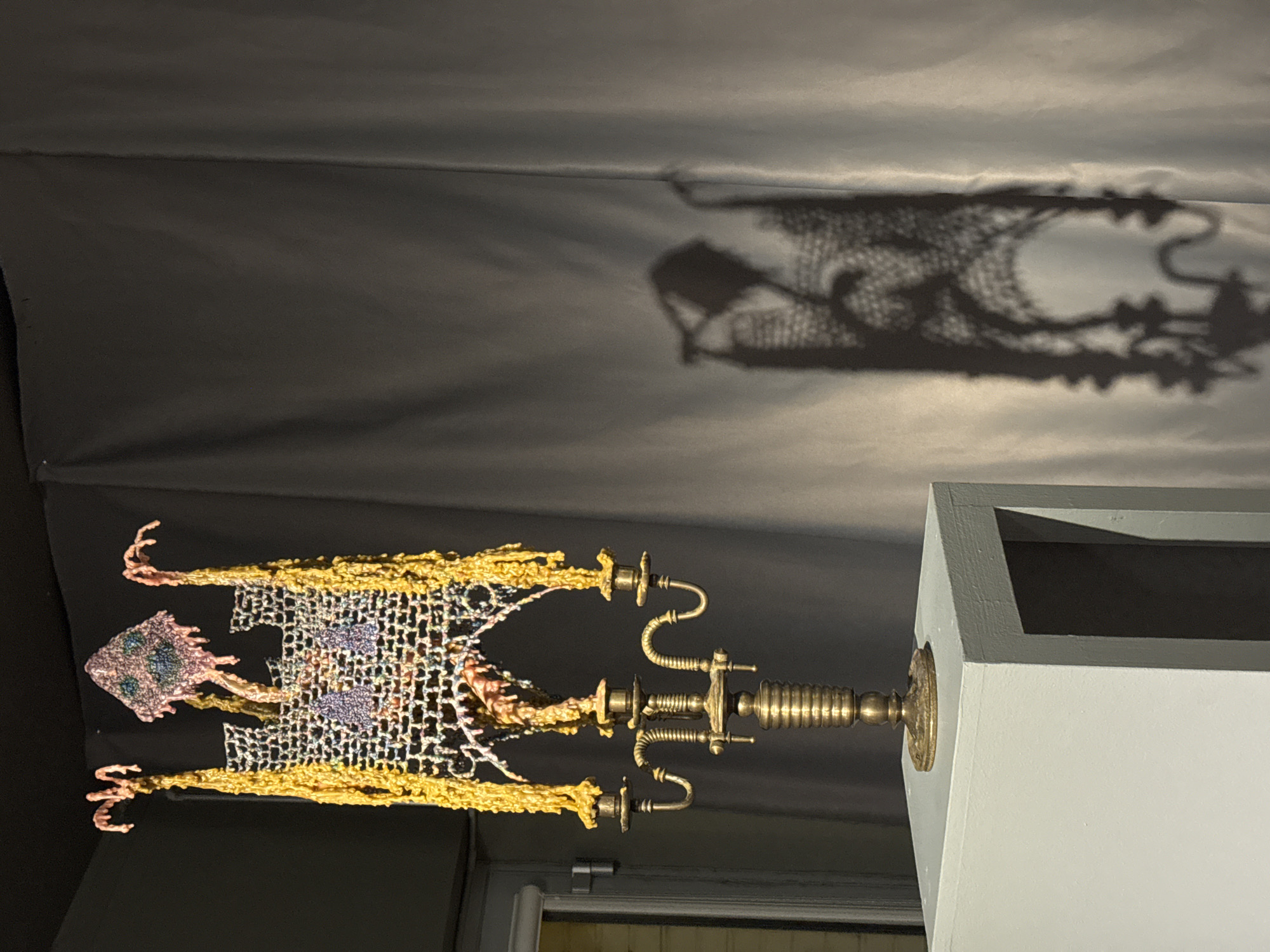 The image shows an ornate, metallic sculpture with a netted structure and decorative extensions resembling dripping gold and coral appendages. It is mounted on a light gray pedestal, positioned against a dark gray backdrop. A distorted shadow of the sculpture is cast on the backdrop, suggesting a light source from the lower right. The sculpture's metallic components appear to be aged or patinated. The environment appears to be an indoor space, possibly a gallery or museum setting. There is no indication that it is located in Moscow, Russia. There is no people, subjects, actions, or interactions.