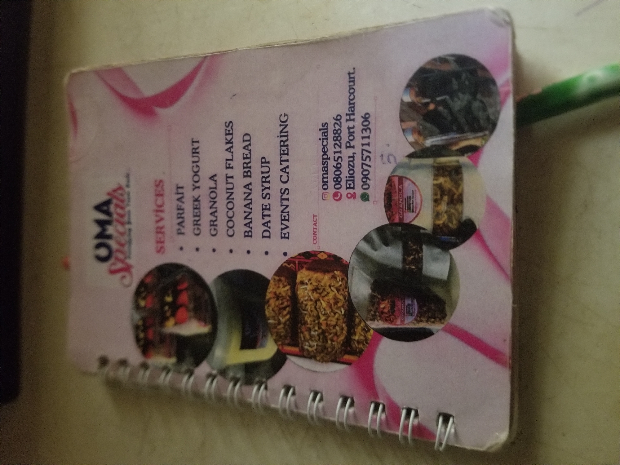 The media file shows a close-up, angled view of a spiral-bound notebook or notepad. The cover features a pink and white design with abstract wavy lines and circular images of food items. The visible text on the cover includes the business name "OMA Specials" and a list of services such as "PARFAIT", "GREEK YOGURT", "GRANOLA", "COCONUT FLAKES", "BANANA BREAD", "DATE SYRUP", and "EVENTS CATERING". There are also contact details, including an Instagram handle "@omaspecials", a phone number "08065128826", an address "Eliozu, Port Harcourt.", and another phone number "09075711306". The circular images depict what appear to be jars or containers of granola and other food items. The notebook is resting on a textured surface, possibly a table, and a green pen is partially visible in the upper right corner. The lighting suggests an indoor setting. The overall impression is of a promotional item for a food-related business, likely based in Port Harcourt, Nigeria.
